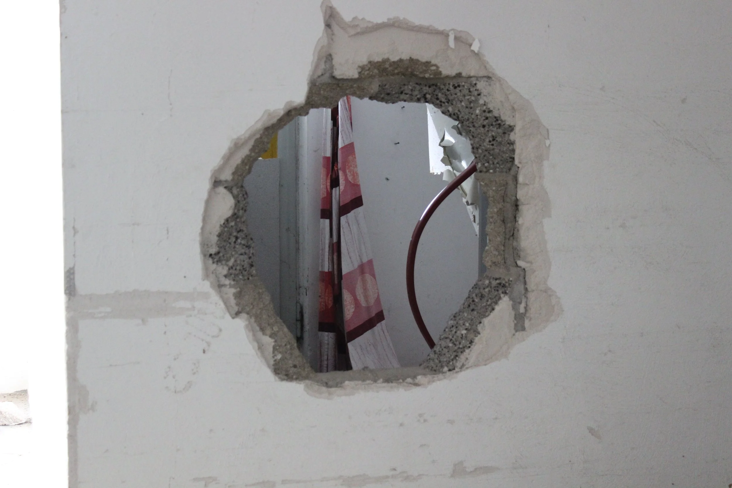 Loch in einer weißen Wand mit sichtbarem Baustoff darunter, Blick durch das Loch auf einen Raum mit einer roten Wasserleitung und einem Vorhang mit roten und weißen Mustern.