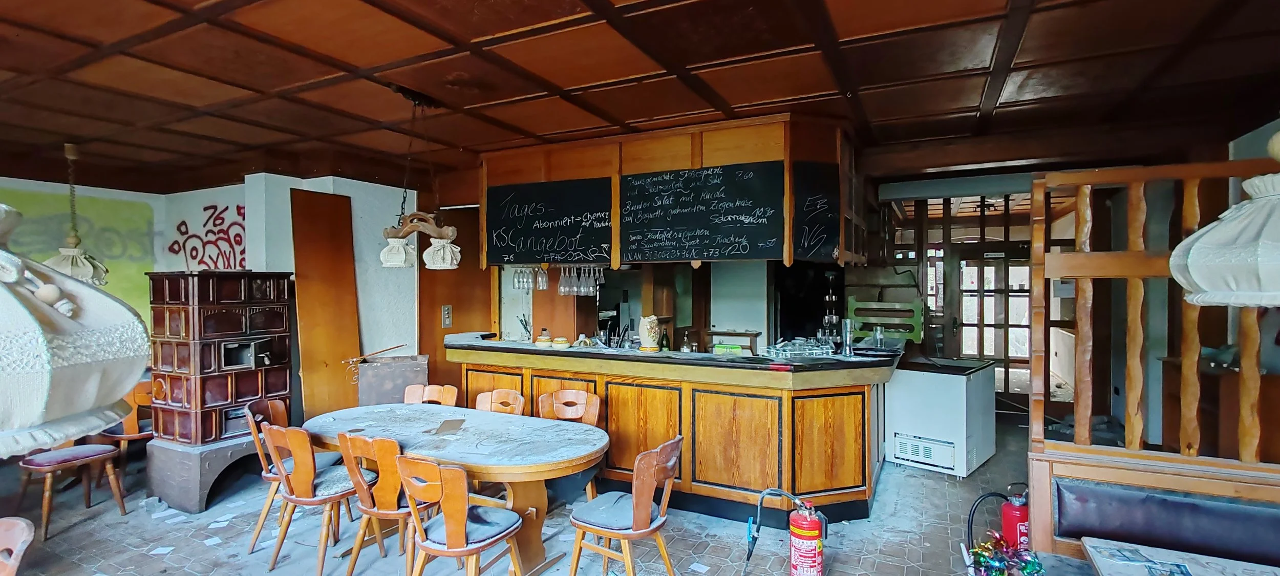 Innenaufnahme eines rustikalen Restaurants mit Holzinterieur, einem runden Esstisch mit acht Stühlen, einer Bar mit Gläsern und schwarzen Tafeln mit Speisekarten.