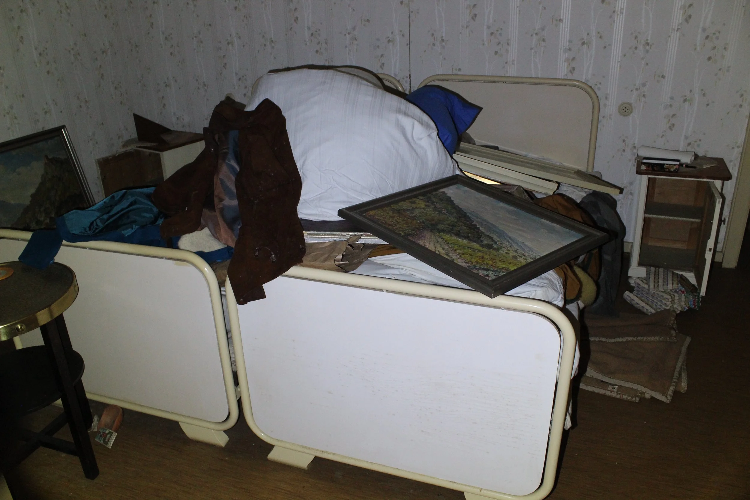 Ein unordentliches Bett mit verstreuten Kleidern und einem Kunstbild auf der Decke in einem Zimmer mit alter Tapetenwand und kleinem Nachttisch.