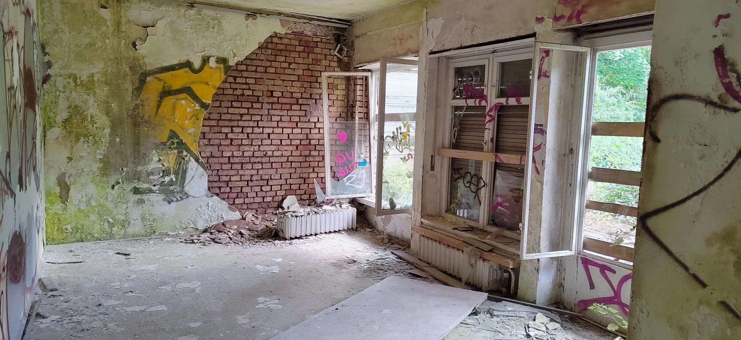 Ein verfallenes, graffitiüberwachtes Zimmer mit offenen Fenstern, teilweise abblätternde Wände und herumliegenden Schutt.