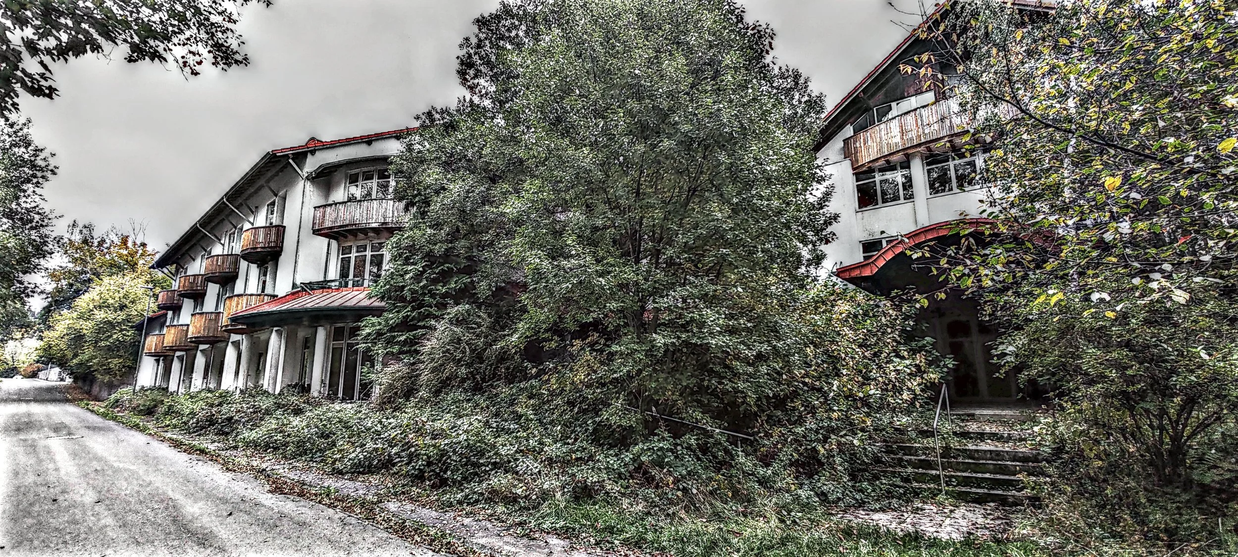 Ein mehrstöckiges Gebäude mit Balkonen, teils verrostete Geländer, umgeben von Bäumen und Sträuchern, vor einer unbefestigten Straße.