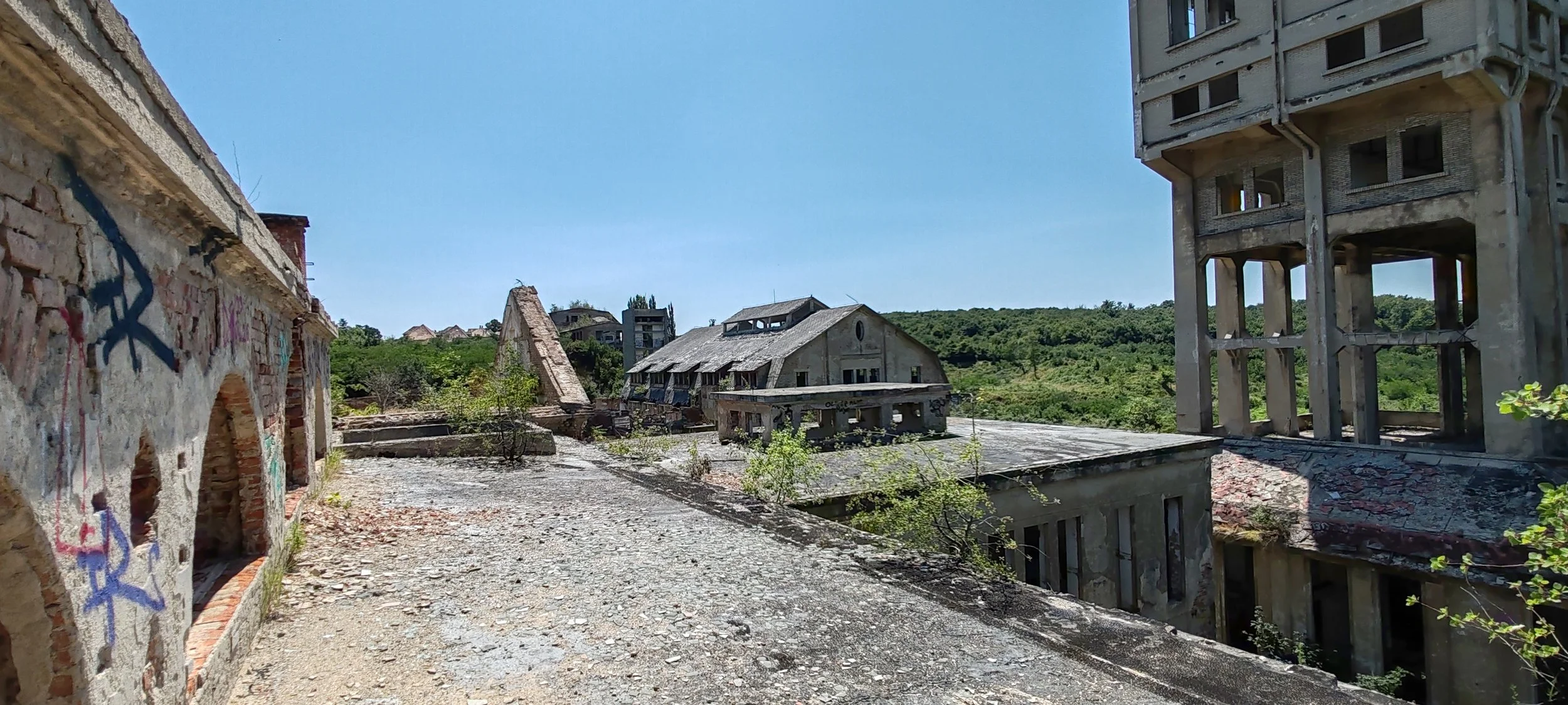 Verfallene Gebäude und Ruinen in einer verlassenen Gegend, umgeben von grünen Hügeln und Bäumen, bei sonnigem Himmel.