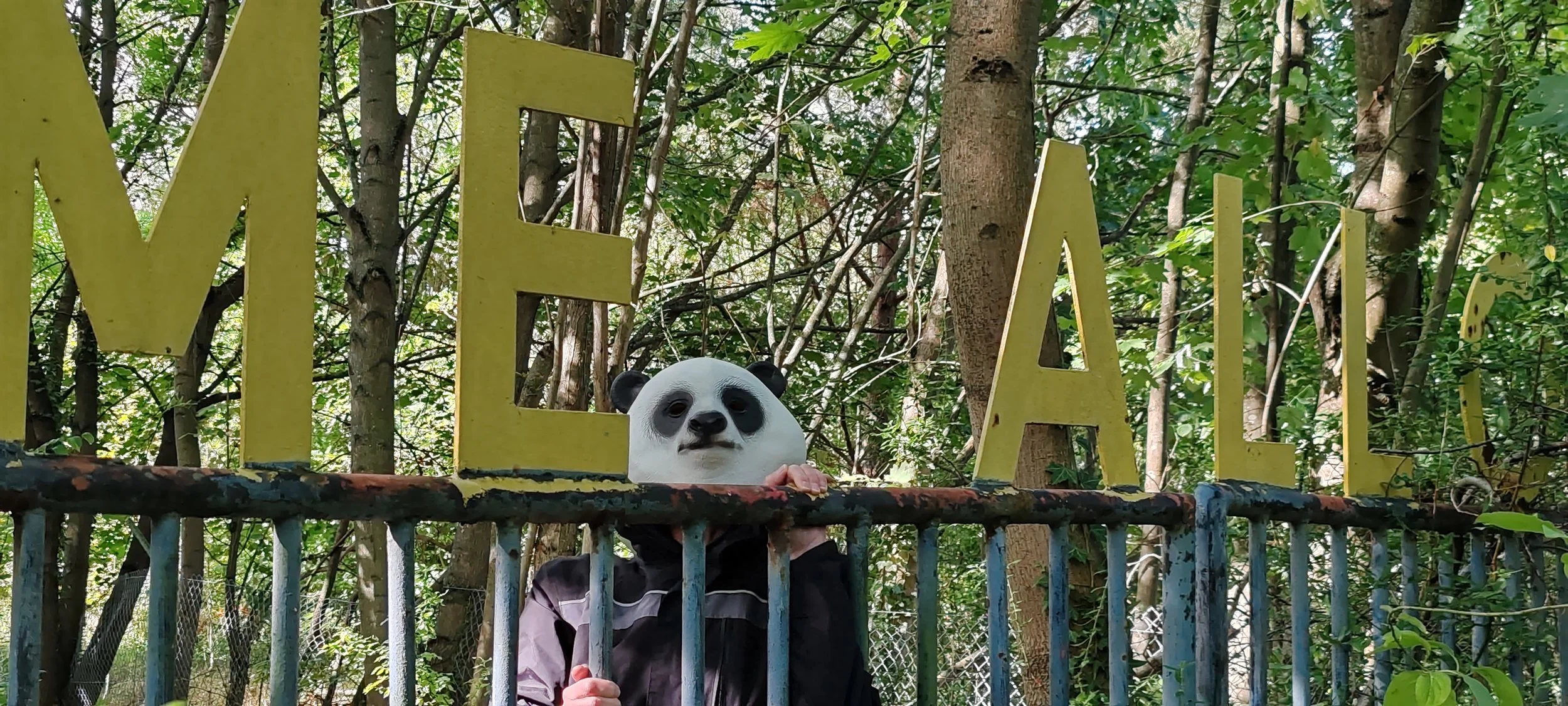 Ein Kind trägt eine Panda-Maske und steht vor einem Zaun im Wald, neben einem gelben Schriftzug, der das Wort »WELT« bildet.