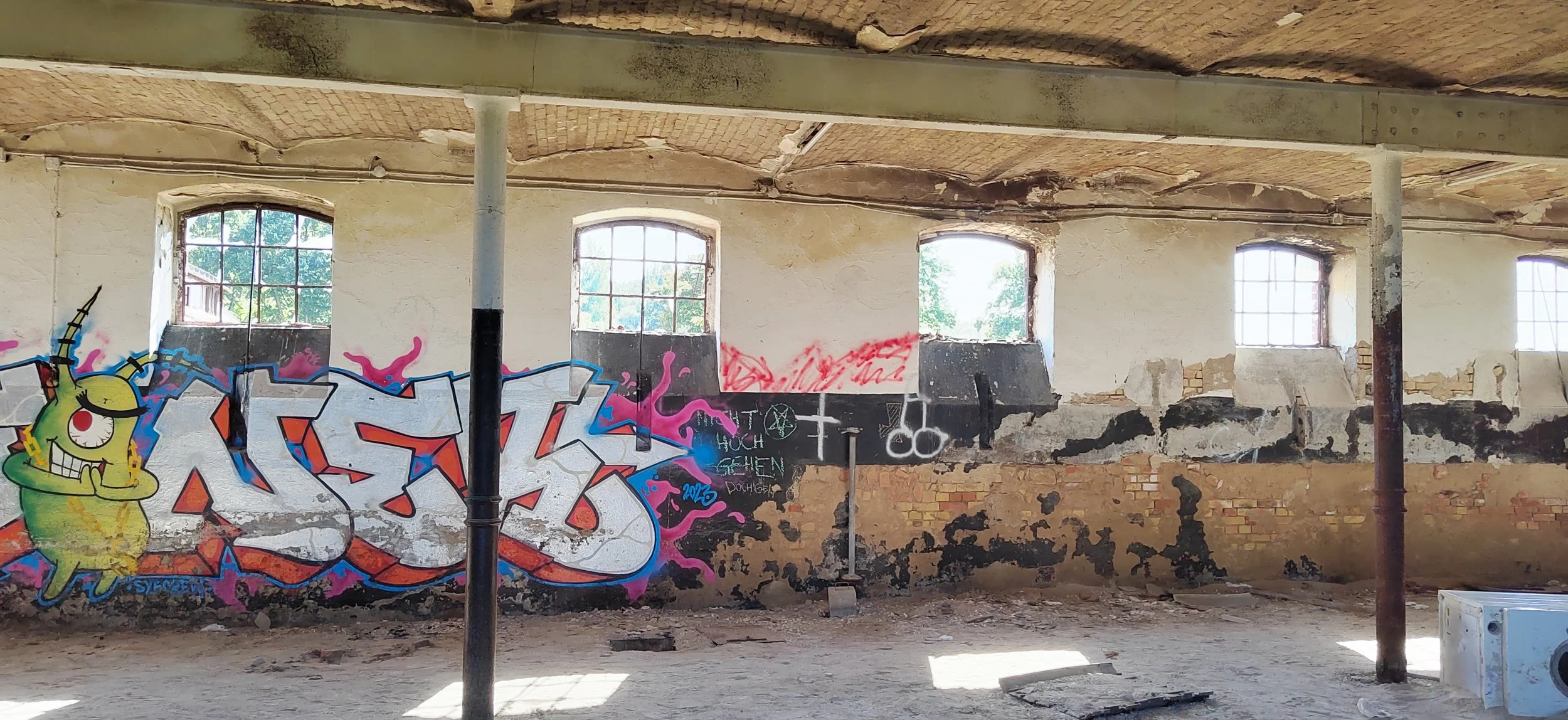 Innenansicht eines verfallenen Gebäudes mit vier großen fenstern, an der Wand Graffiti, das ein grünliches Monster mit einem Horn und geschlossenen Augen zeigt, sowie andere farbige Schriftzüge und Symbole.