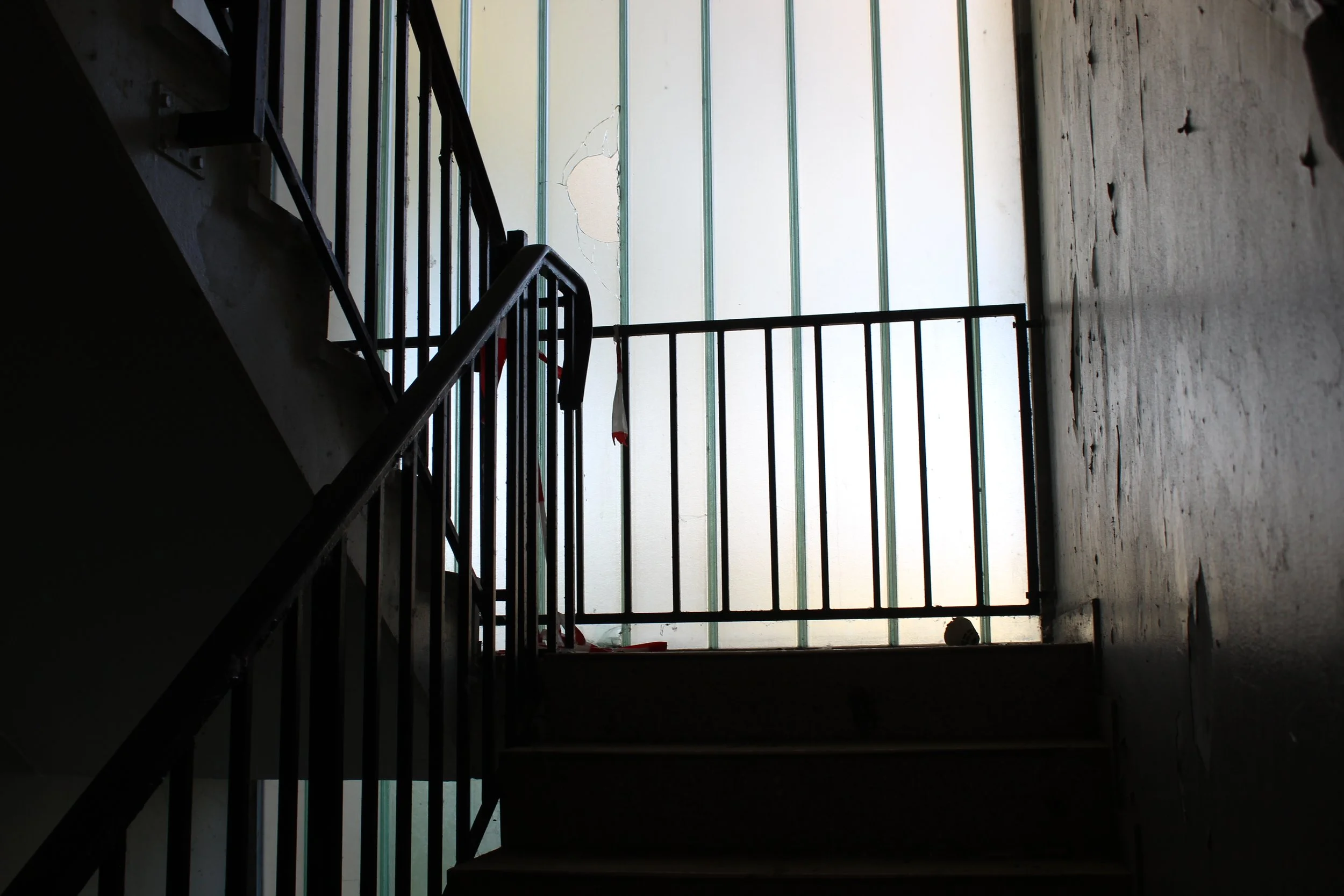 Blick von unten auf eine dunkle Treppe mit Schwarzstahlgeländer, vor einer Wand mit Glasfenstern, im oberen Bereich eines Gebäudes.
