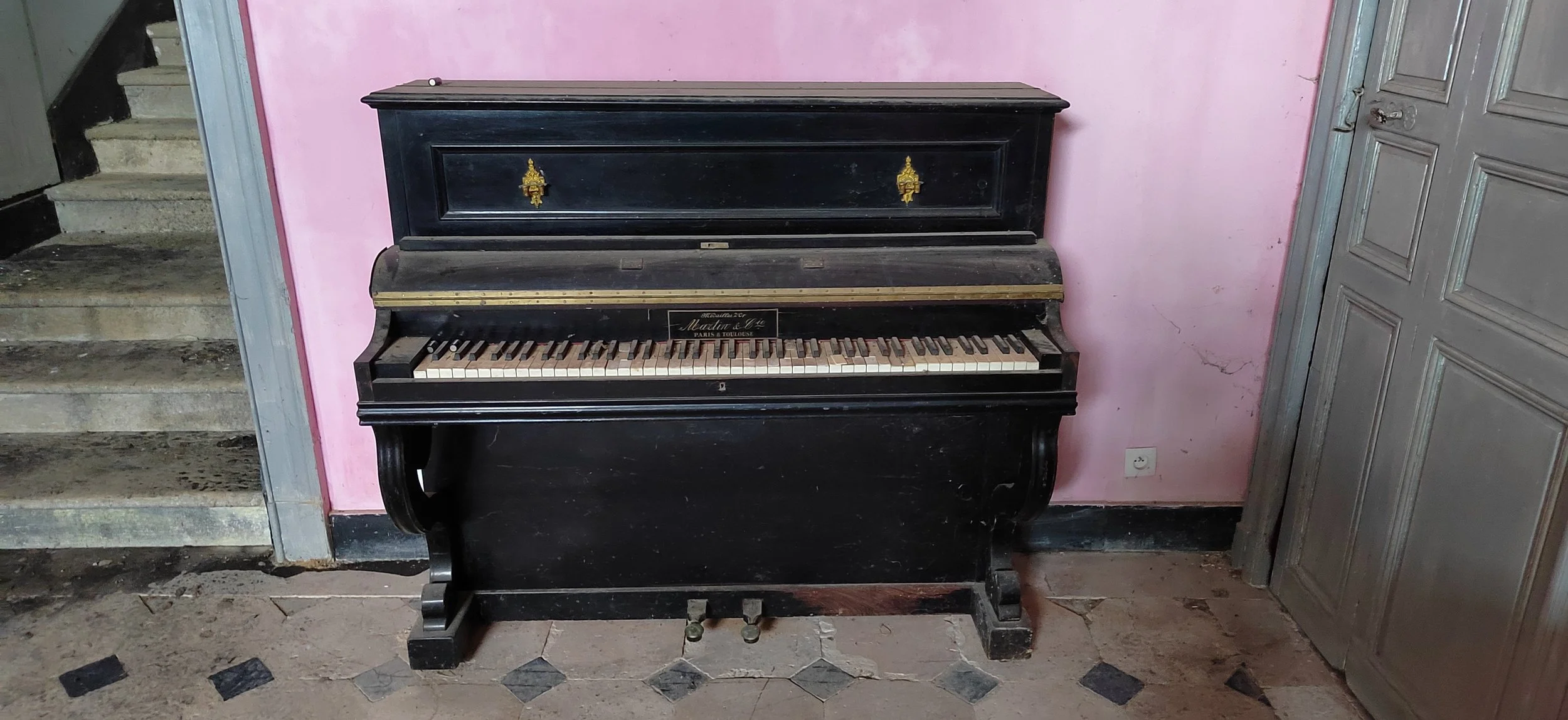 Alter schwarzes Klavier mit missgestalteten Tasten, aufgestellt in einem Raum mit rosa Wand und Steinstufen auf der linken Seite.