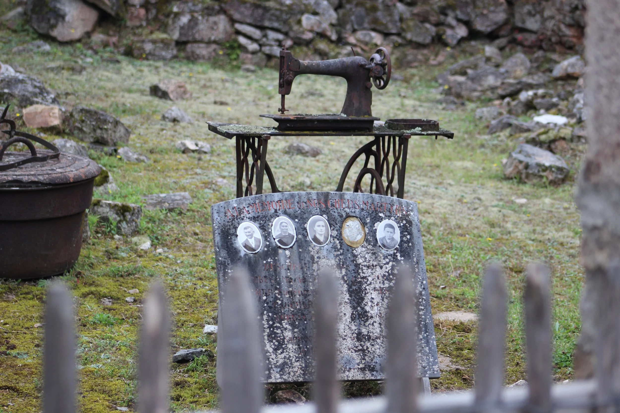 Ein antikes Handmaschinen-Nähmaschine auf einem Holzgestell steht auf einer Wiese mit Steinen. Vor der Maschine befindet sich eine Gedenktafel mit schwarzen und weißen Porträtfotos und deutschen Texten, die nach einem Krieg oder Konflikt zu lohnen sc