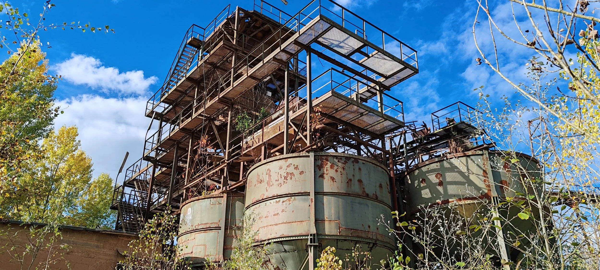 Ein alter, rostiger Industriekomplex mit mehreren Ebenen und Leitern, umgeben von Bäumen, vor blauem Himmel.