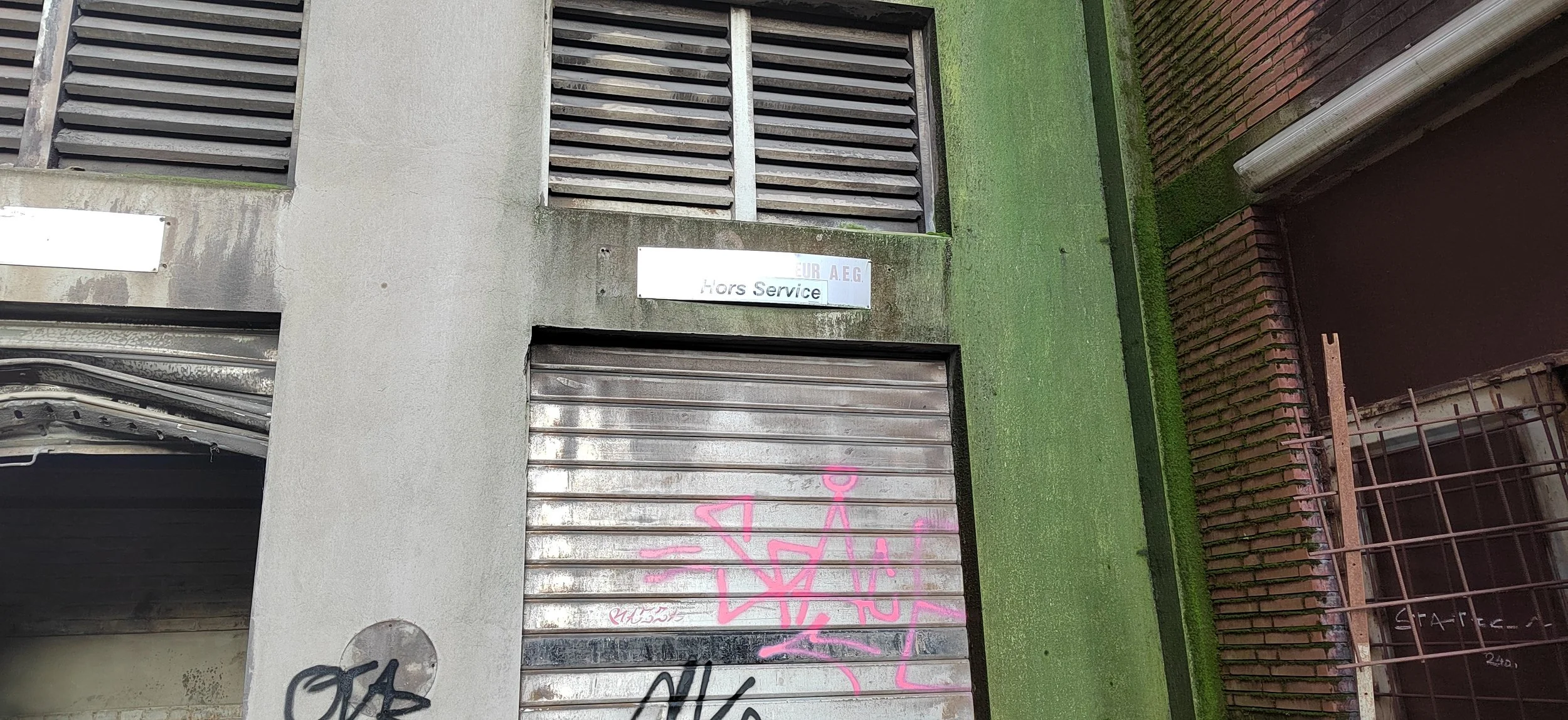 Ein geschlossenes Garagentor mit Graffiti in pink Farbe und einer grauen Wand mit einem schwarzen Graffiti. Über dem Garagentor ist ein silbernes Schild mit der Aufschrift "Hors Service".