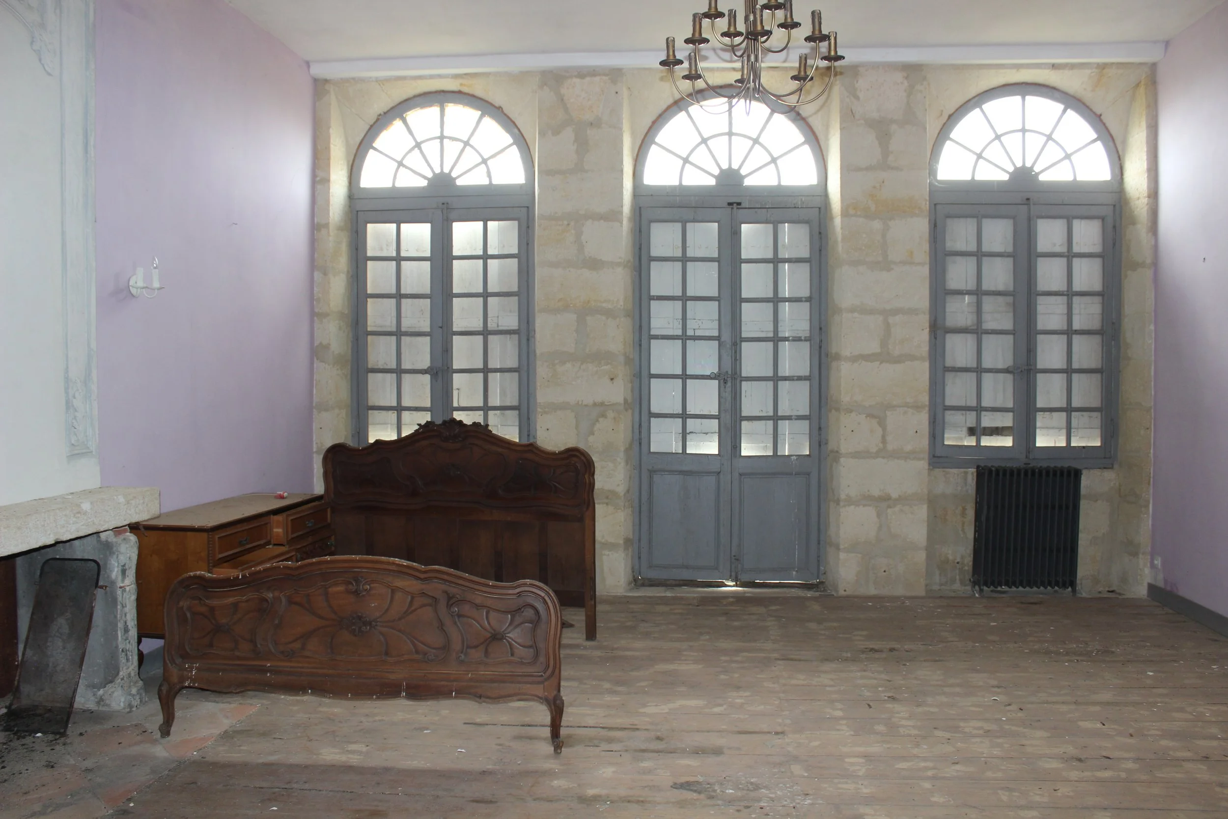 Ein leeres Wohnzimmer mit drei großen fensterartigen Türen mit Bögen, altem Holzparkettboden, einer alten Holzkommode und einem Kronleuchter an der Decke.