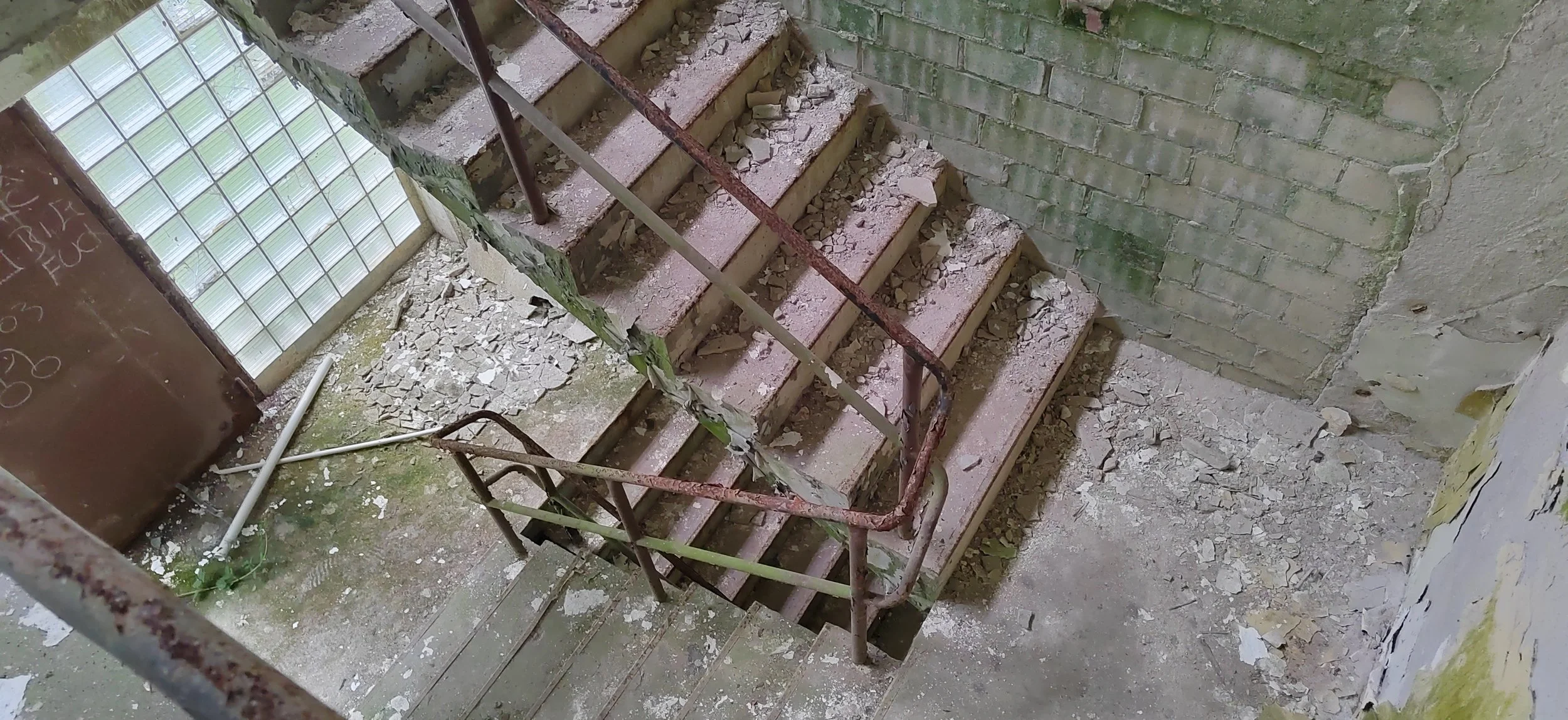Verfallene Treppe in einem alten Gebäude, mit zerbrochenem Beton und rostigem Geländer, an einer Wand aus Ziegelsteinen.