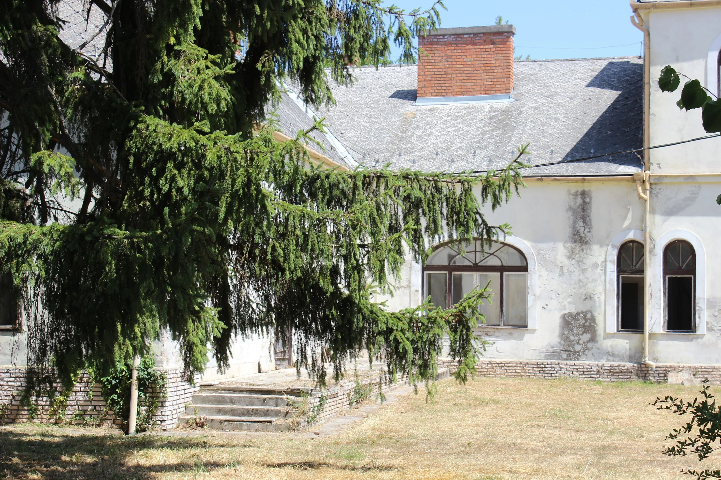 Ein altes, weißes Gebäude mit verzierten Fenstern und einem roten Schornstein, teilweise verdeckt von einer großen Tannenbaum. Der Hof ist überwiegend trockenes Gras, es gibt einige Stufen, die zum Eingangsbereich führen.