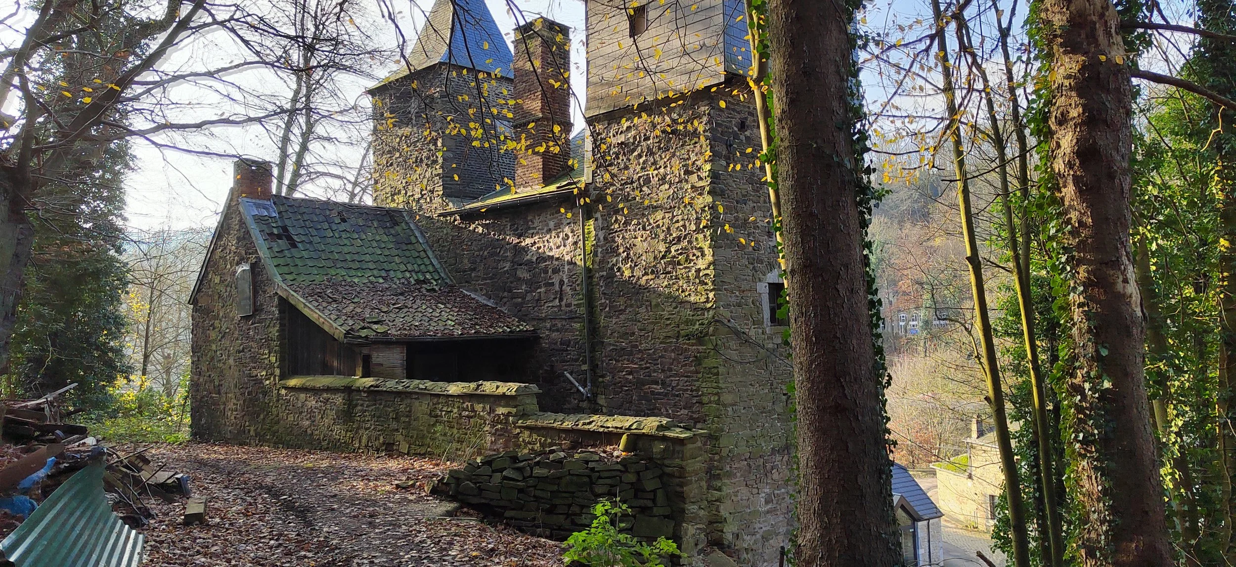 Ein altes verfallenes Steinhaus im Wald, umgeben von Bäumen mit gelben und grünen Blättern, mit einem verschmutzten Dach und einer unebenen Erde im Vordergrund.