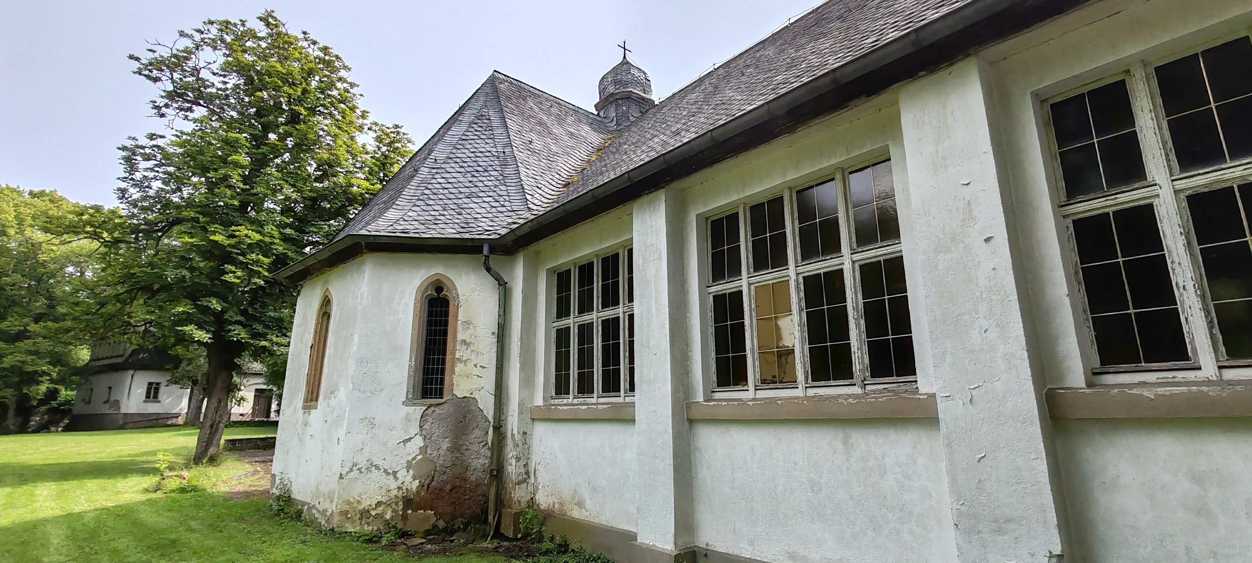 Alte Kirche mit weißen Wänden, großen Fenstern und einem spitzen Dach, umgeben von grünem Rasen und Bäumen