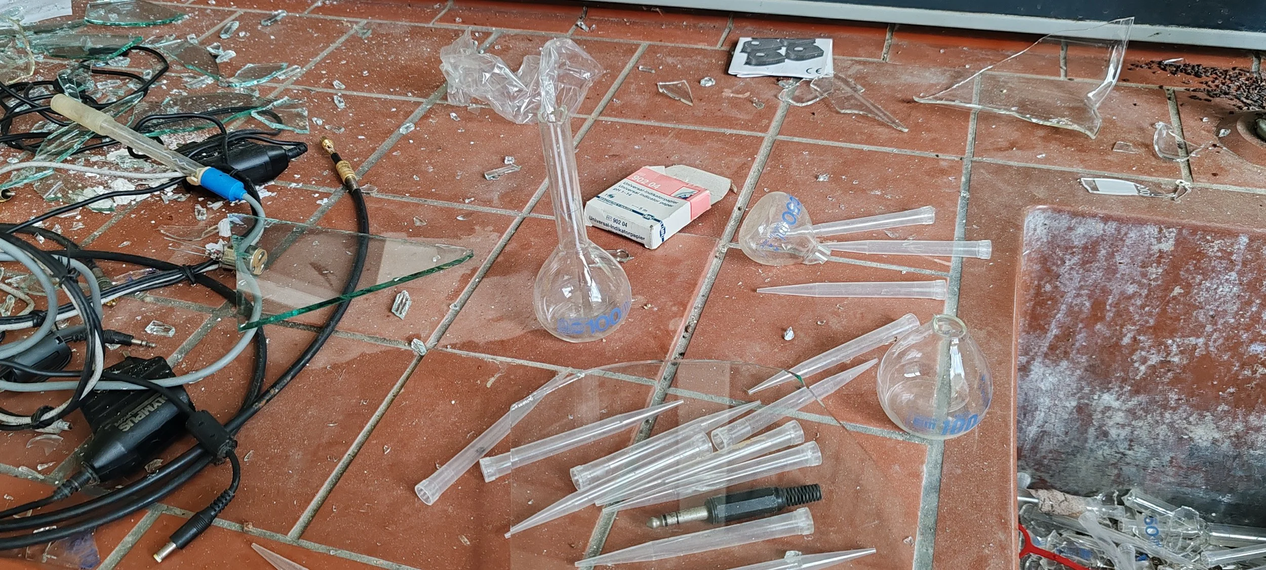 Zerbrochenes Labor- oder wissenschaftliches Zubehör, darunter Glasröhren, Messzylinder, Kabel, und Verpackungen, auf einem Fliesenboden.