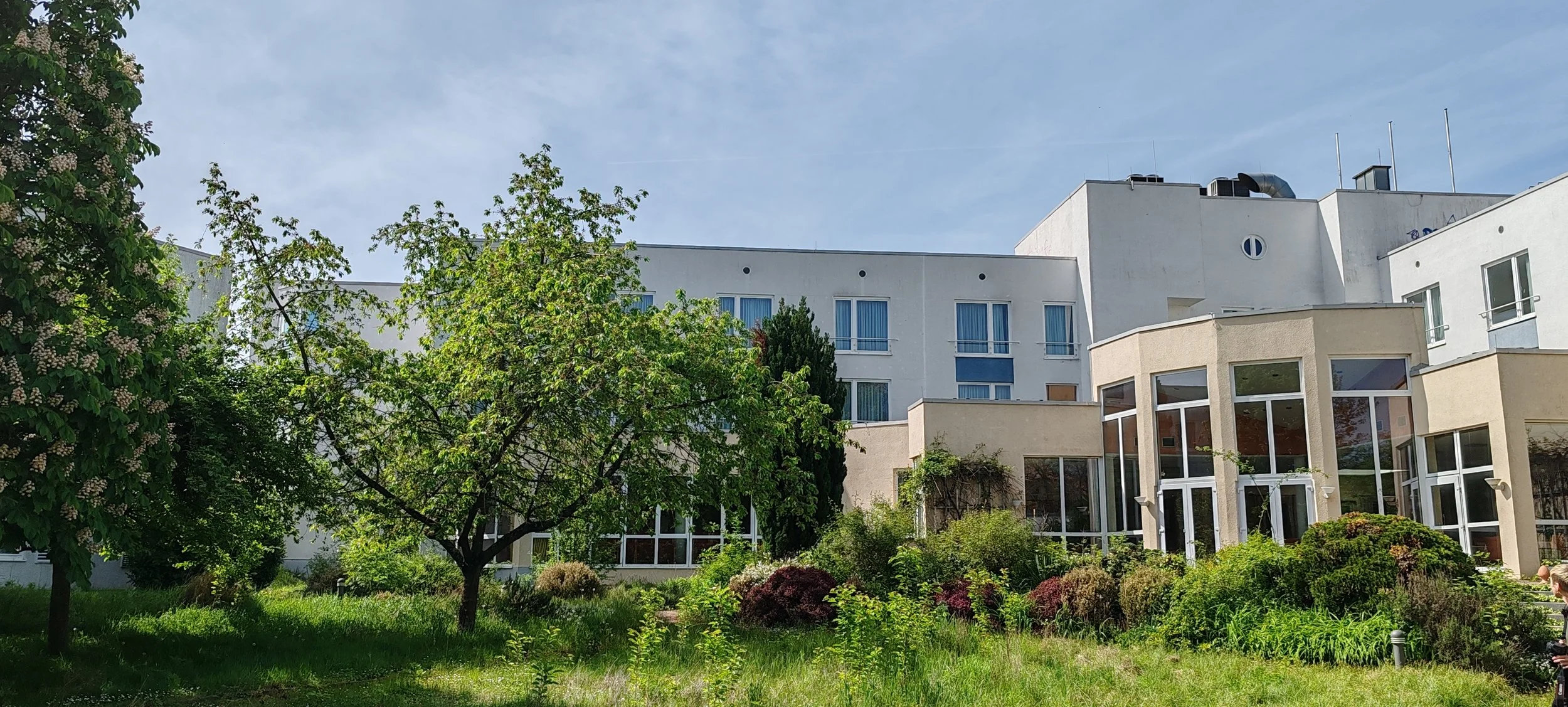 Moderner Gebäudekomplex mit großen Fenstern, umgeben von einem grünen Garten mit Bäumen und Sträuchern bei sonnigem Himmel.