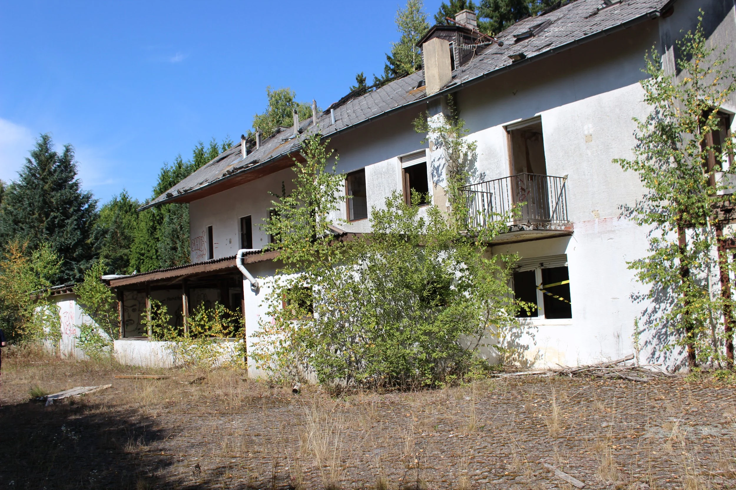 Ein verfallenes, weißes, mehrstöckiges Gebäude mit zerstörten Fenstern und einer verrosteten Balustrade, umgeben von toten Pflanzen und Bäumen, mit einem blauen Himmel im Hintergrund.