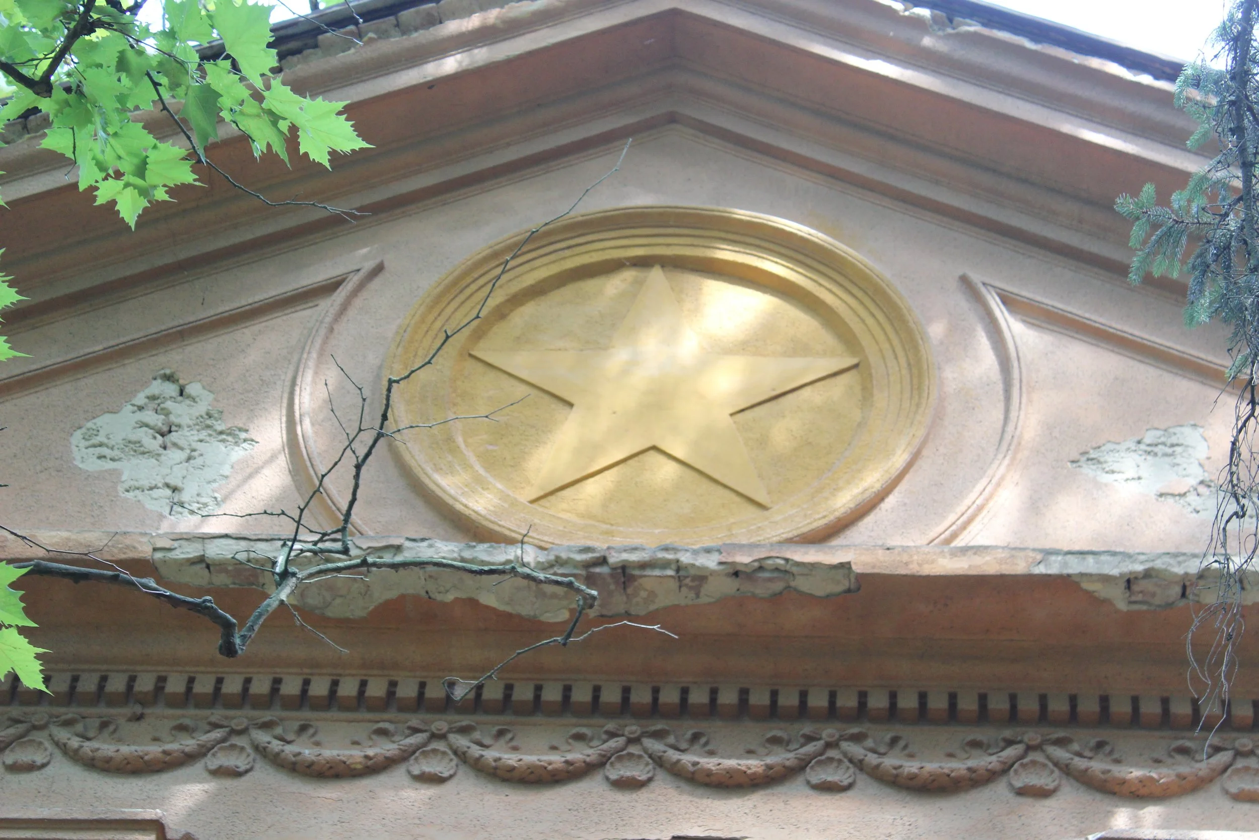 Fassade eines Gebäudes mit goldener Sterndekor im oberen Bereich, umgeben von grünen Blättern und Zweigen.