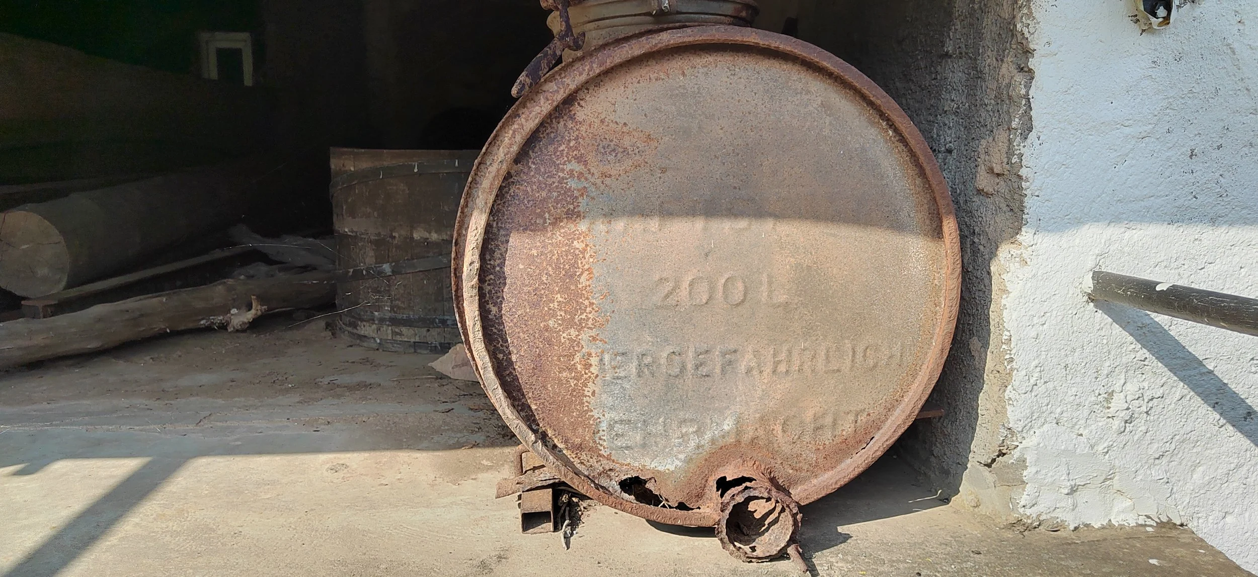 Ein rostiger Metallbehälter mit der Aufschrift 'TÜV 102' steht vor einer weißen Wand. Im Hintergrund befinden sich Holzstämme und andere Metallteile.