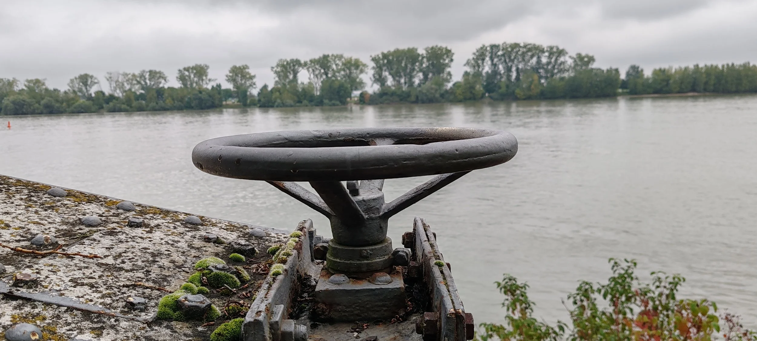 Ein metallischer Anker in der Nähe eines Flusses, umgeben von Mauerwerk mit Moos und kleinen Pflanzen, bei bewölktem Himmel.