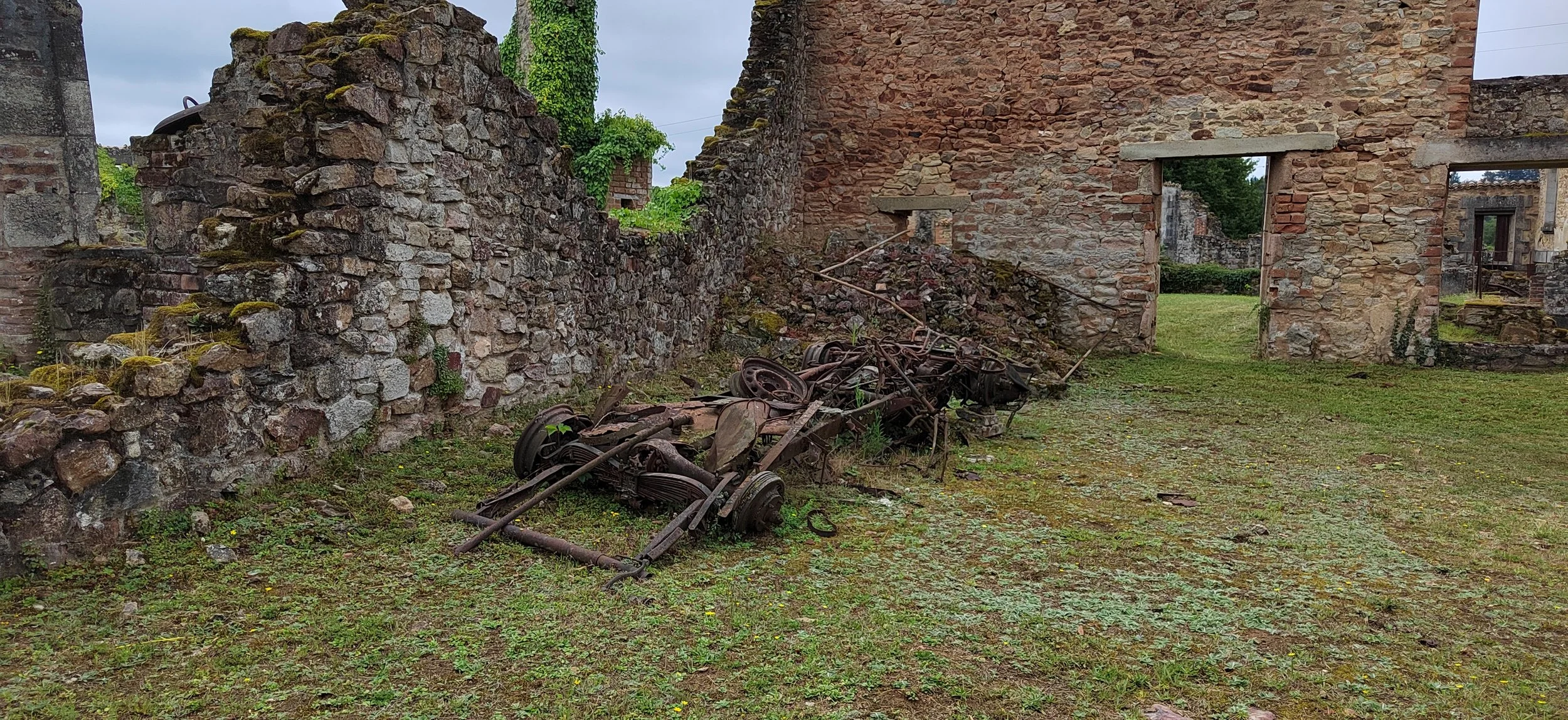 Verrostete, alte landwirtschaftliche Geräte liegen auf dem Boden vor einer alten Mauer aus Ziegelsteinen in einer grünen, bewachsenen Umgebung, vermutlich in einer verlassenen Ruine.