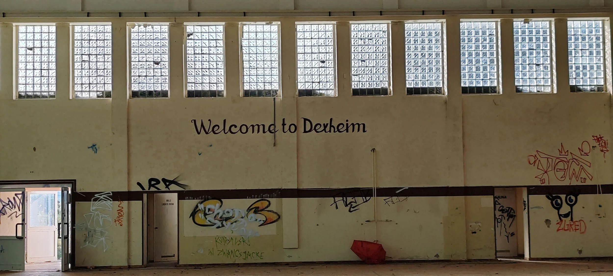 Innenraum eines verlassenen Gebäudes mit einem Graffiti-Wandbild mit dem Schriftzug 'Welcome to Dehem' und mehreren Fenstern aus Glasblöcken an der Decke.