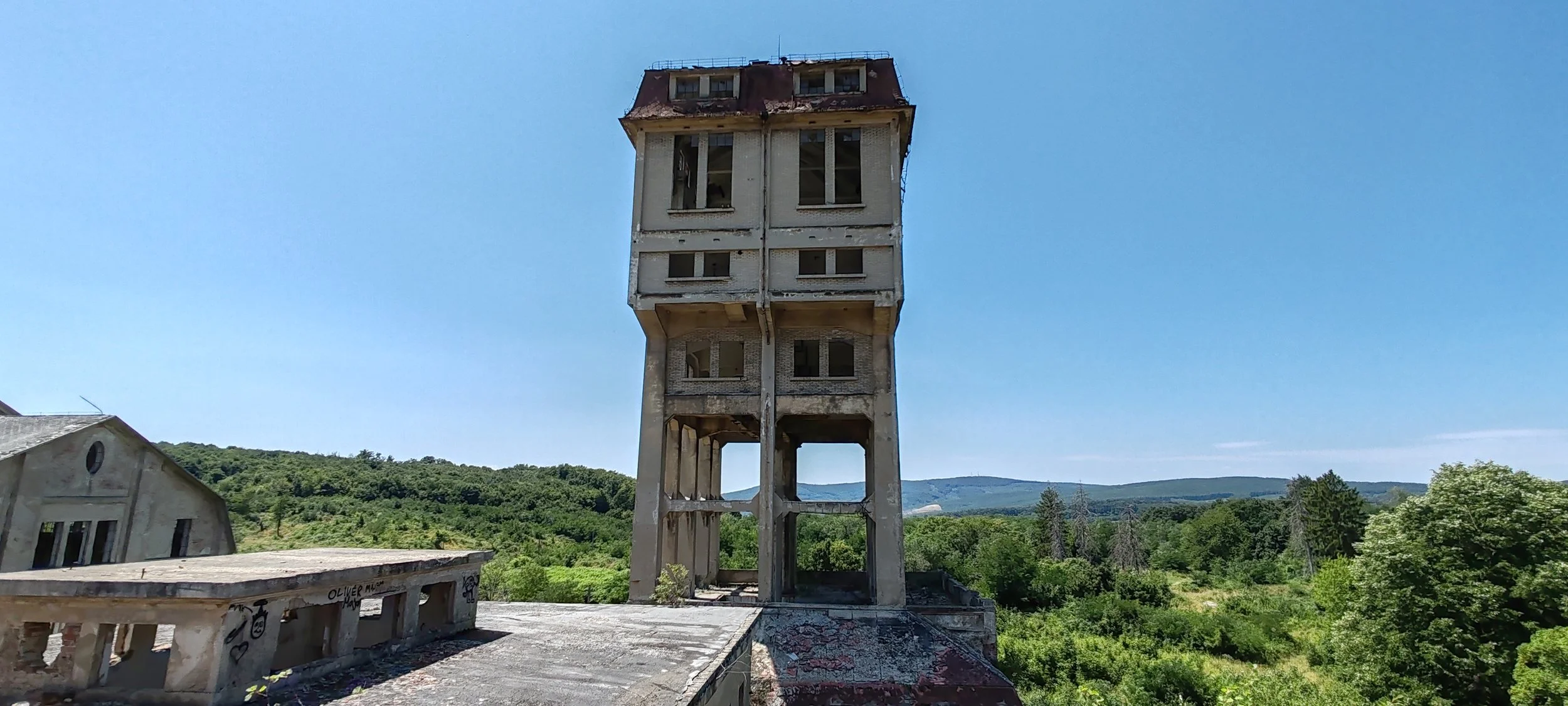 Verfallene hohe Betonturm mit mehreren Fenstern, umgeben von grünem Wald und Hügeln unter blauem Himmel.