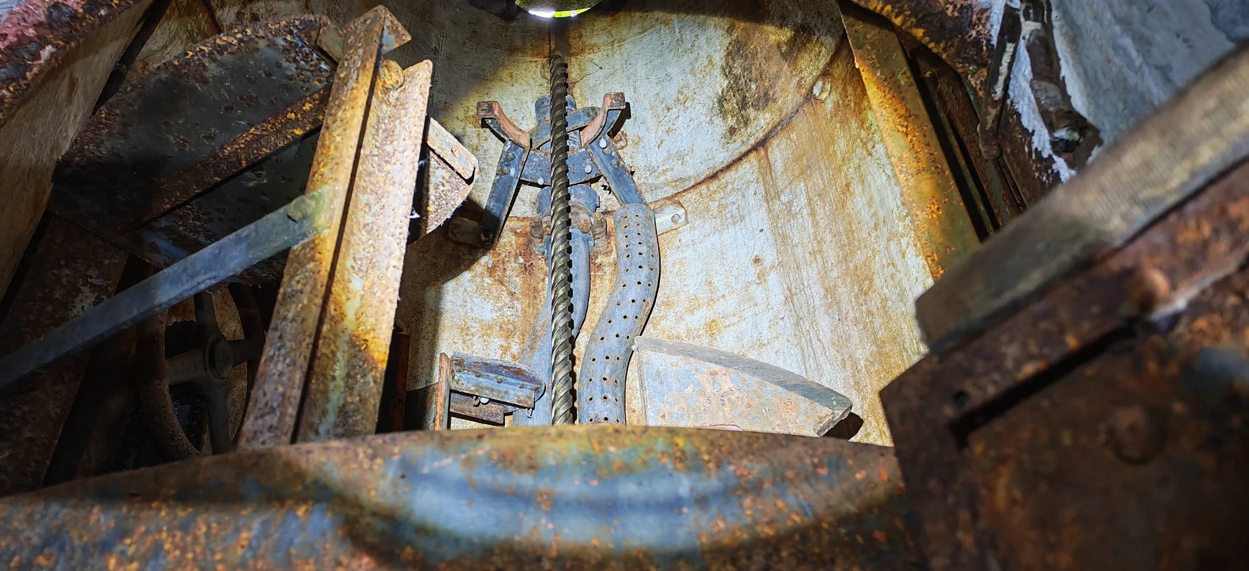 Blick in eine rostige, hohle metallische Struktur mit mechanischen Teilen, darunter eine spiralige Schraube, Metallstäbe und übliche Teile, die auf eine alte, verrostete Maschine oder einen technischen Aufbau hinweisen.