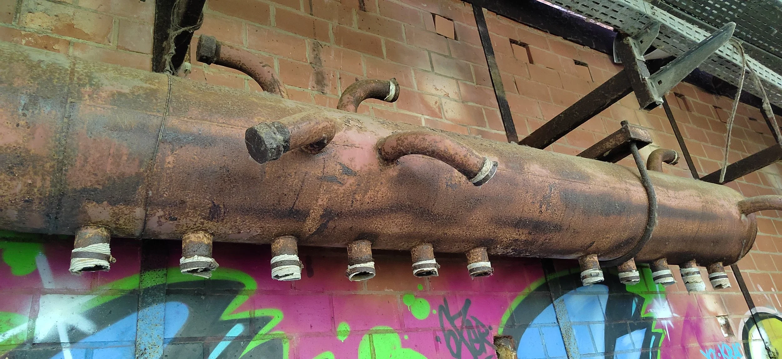 Ein verrosteter, alter Industrietank mit mehreren abgewinkelten Rohren, auf einer bunten, spraygemalten Wand und einer metallenen Treppe im Hintergrund.