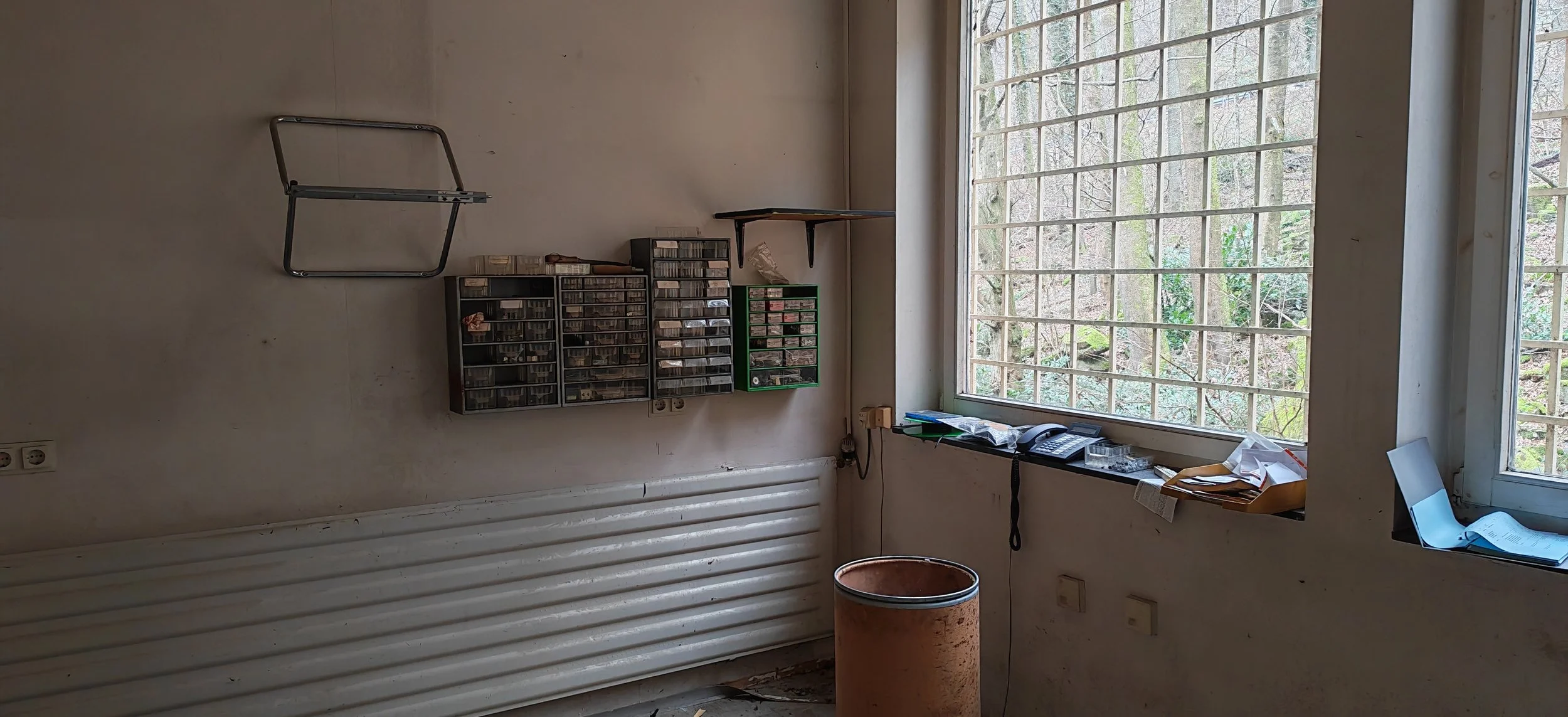 Ein unordentliches Büro mit großen Fenstern, auf denen Gitternetzstäbe sind, und einer Wand mit mehreren kleinen Aufbewahrungsboxen, einem Telefon und Papieren auf der Fensterbank.