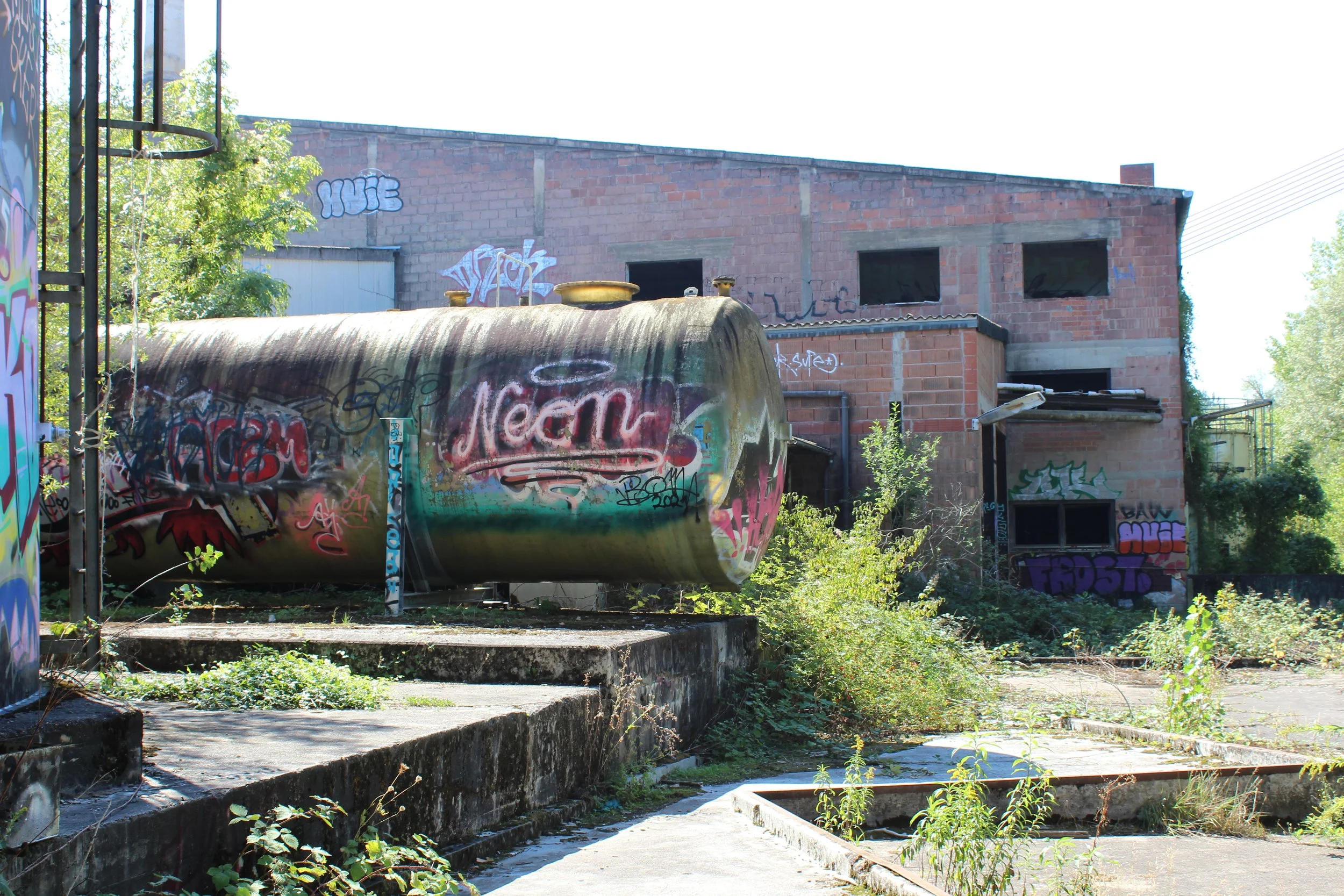 Verfallene, graffiti bewachsene Industrieanlage mit einer großen, mit Graffiti bedeckten Tanks und einer bröckelnden Gebäude im Hintergrund, umgeben von Pflanzen und Überwuchs.