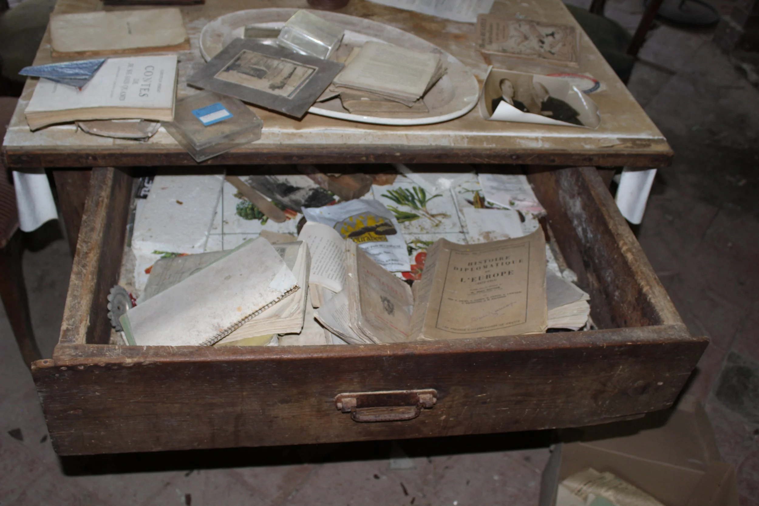 Ein alter, schmutziger Holzschreibtisch mit vielen aufgeschlagenen und verstaubten alten Büchern, Papieren und Fotografien darauf. Der Schreibtisch ist in einem verfallenen Zustand.