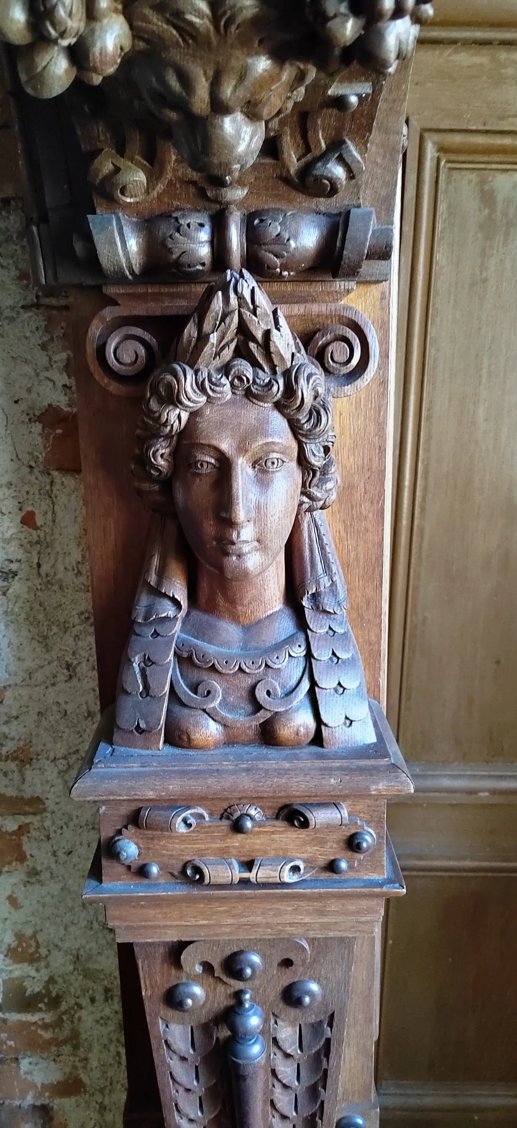 Holzskulptur eines mythologischen Wesens mit einem menschlichen Kopf, umgeben von barocken Verzierungen. Holzrahmen im Hintergrund.