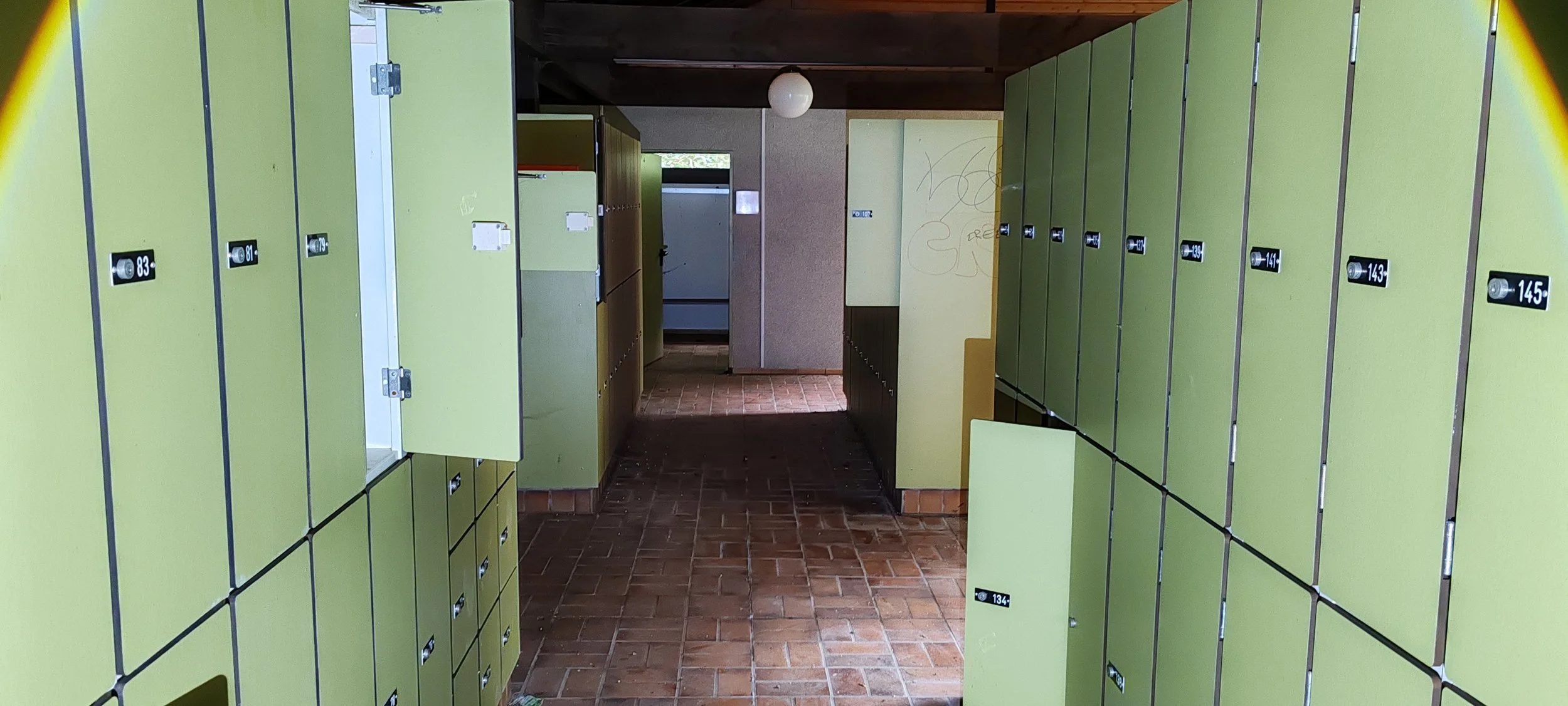 Schließfächer in einer öffentlichen Einrichtung, grüne Farbe, nummeriert, im Flur eines Gebäudes mit Friesenpflasterboden, im Hintergrund eine Tür und eine Wand mit Graffiti.