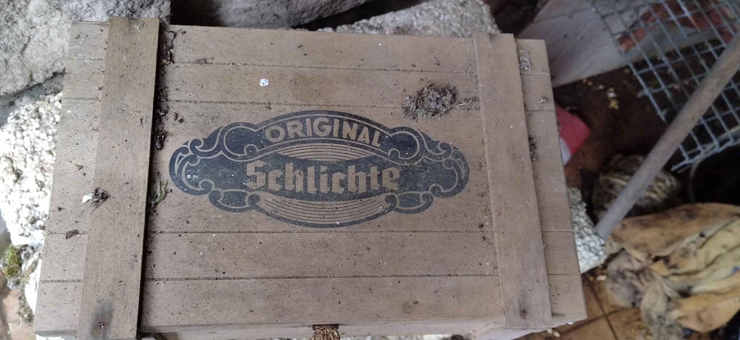 Holzkiste mit dem Aufdruck "Original Schlichte" in Schwarz. Die Kiste liegt auf einem Boden mit Kies und ist teilweise von anderen Baumaterialien umgeben.