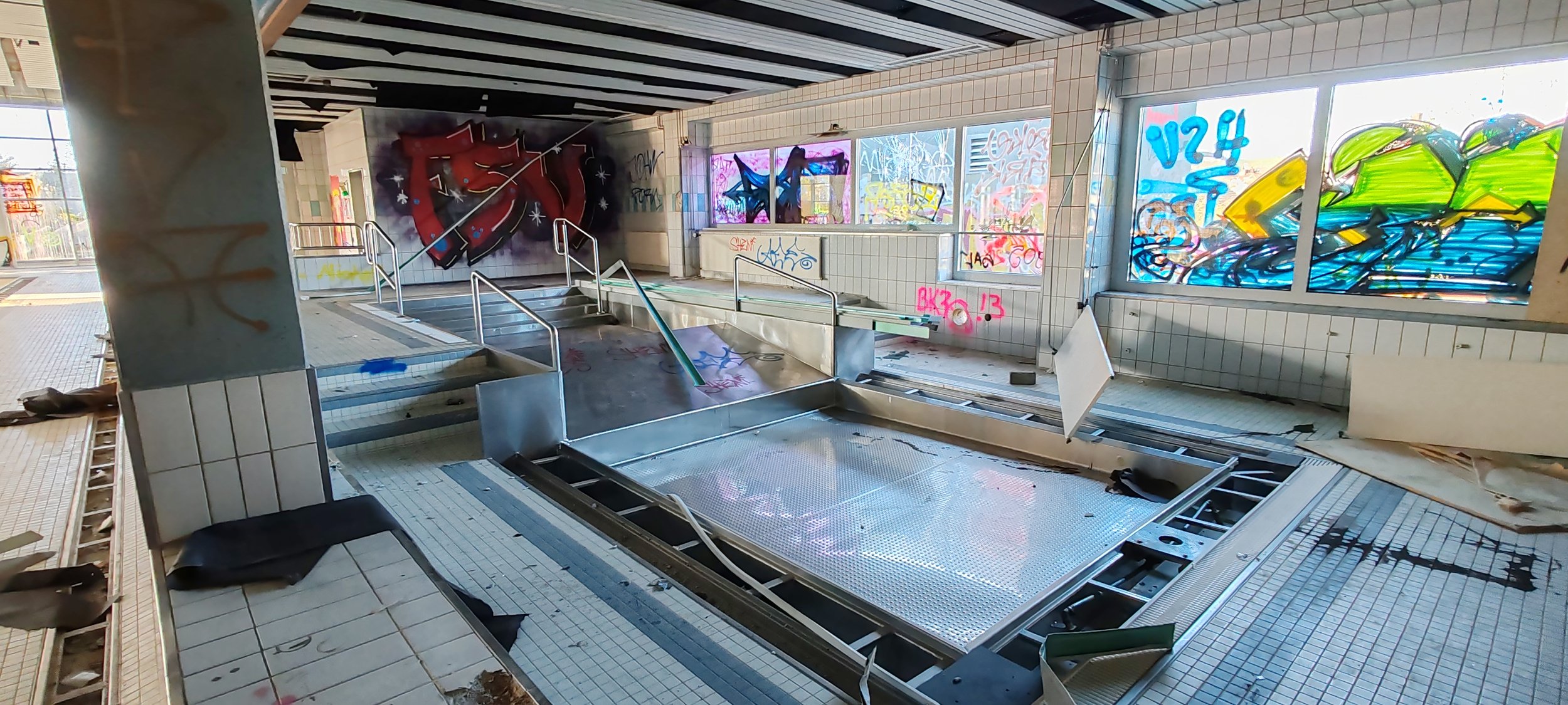Ein verfallenes Schwimmbad mit Graffiti an den Wänden, zerbrochenen Fenstern und Müll auf dem Boden.