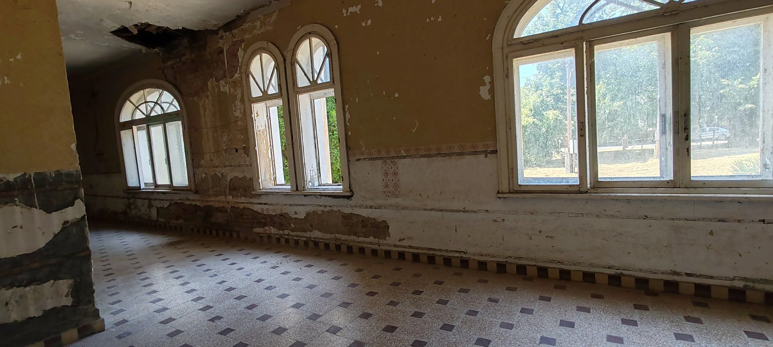 Innenansicht eines alten, renovierungsbedürftigen Raumes mit vier hohen Fenstern, abgenutzten Wänden und Mosaikboden.