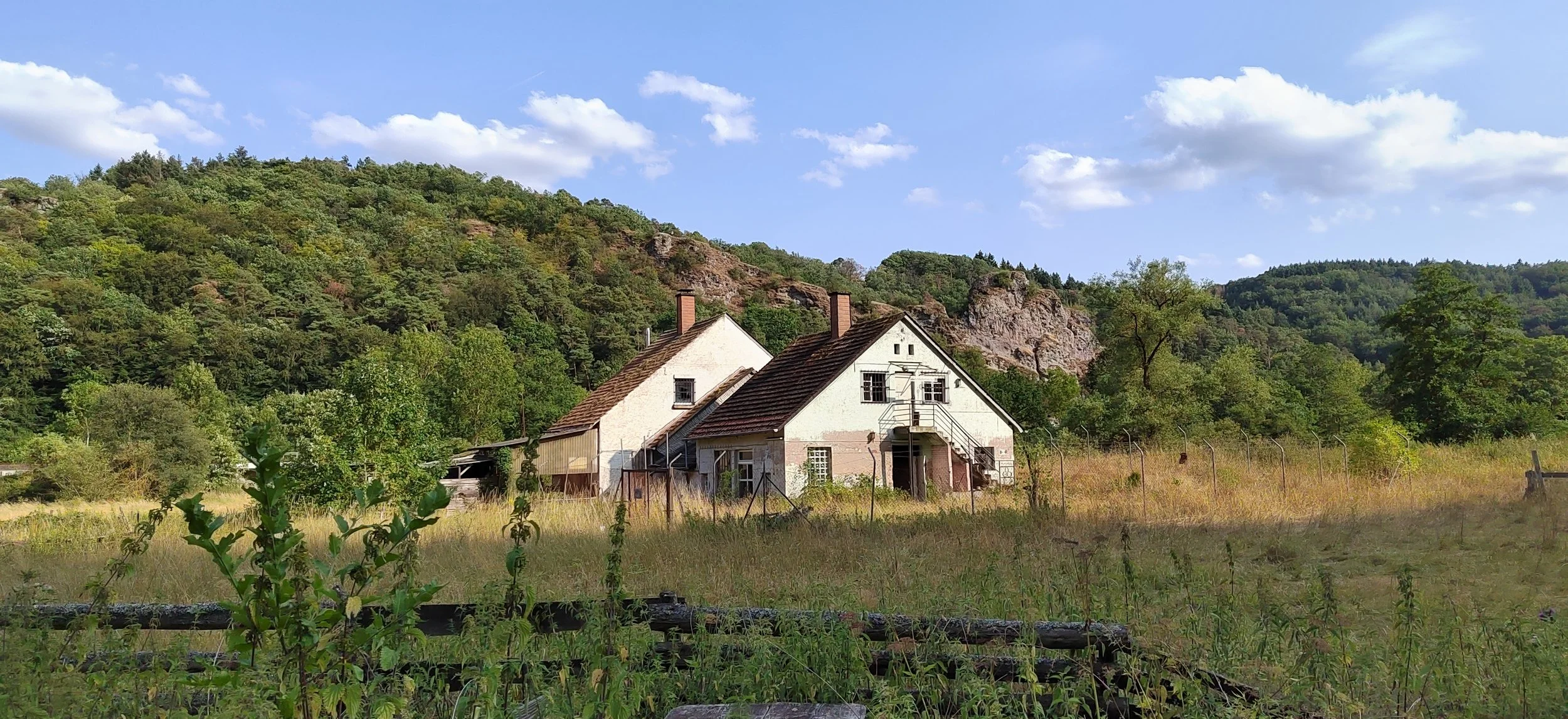Zwei alte Häuser in einer Wiesenlandschaft mit Hügeln und Bäumen im Hintergrund, unter einem blauen Himmel mit Wolken