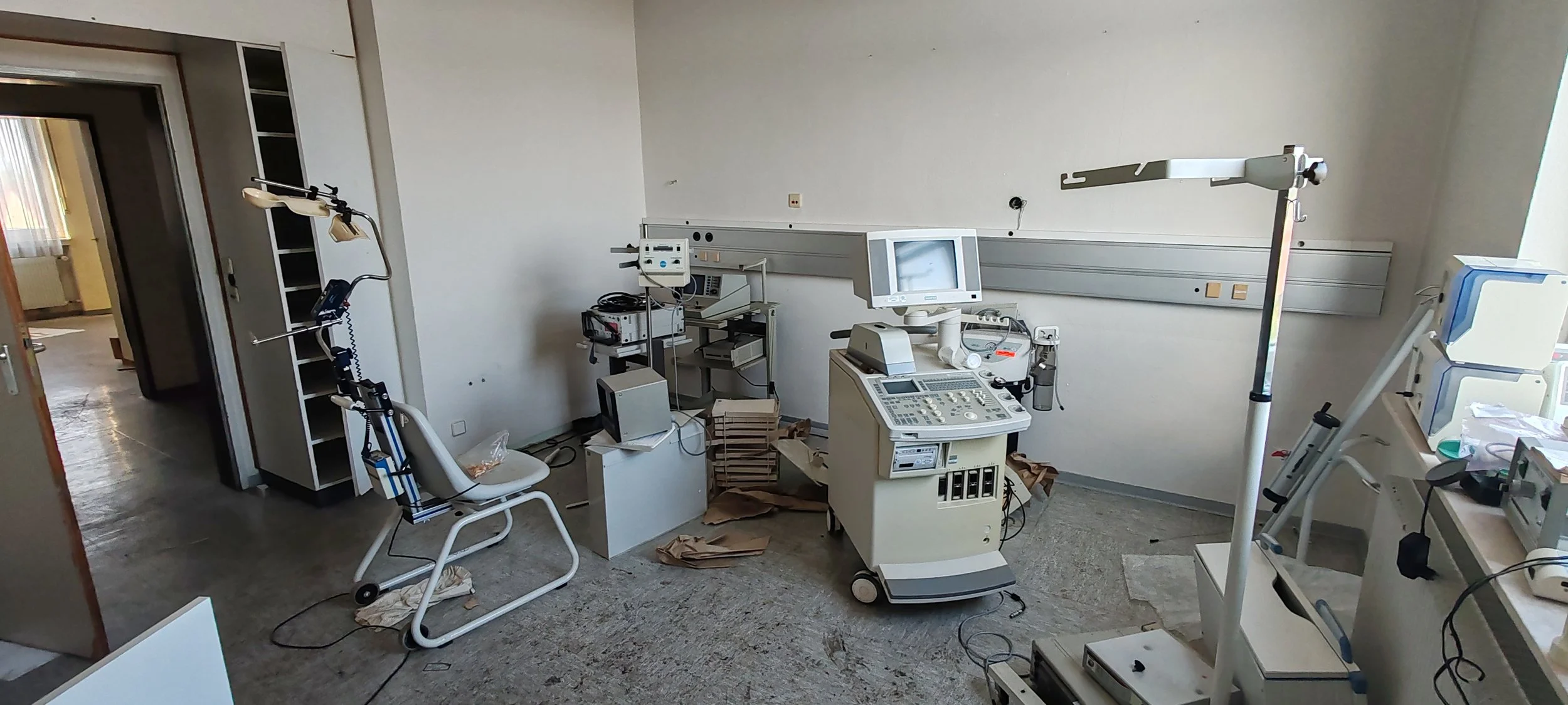 Das Bild zeigt ein verlassene und unordentliche medizinische Station mit veralteter medizinischer Ausrüstung und Geräten, darunter ein Herzmonitorterminal, ein OP-Licht, eine Krankenliege, und diverses medizinisches Zubehör.