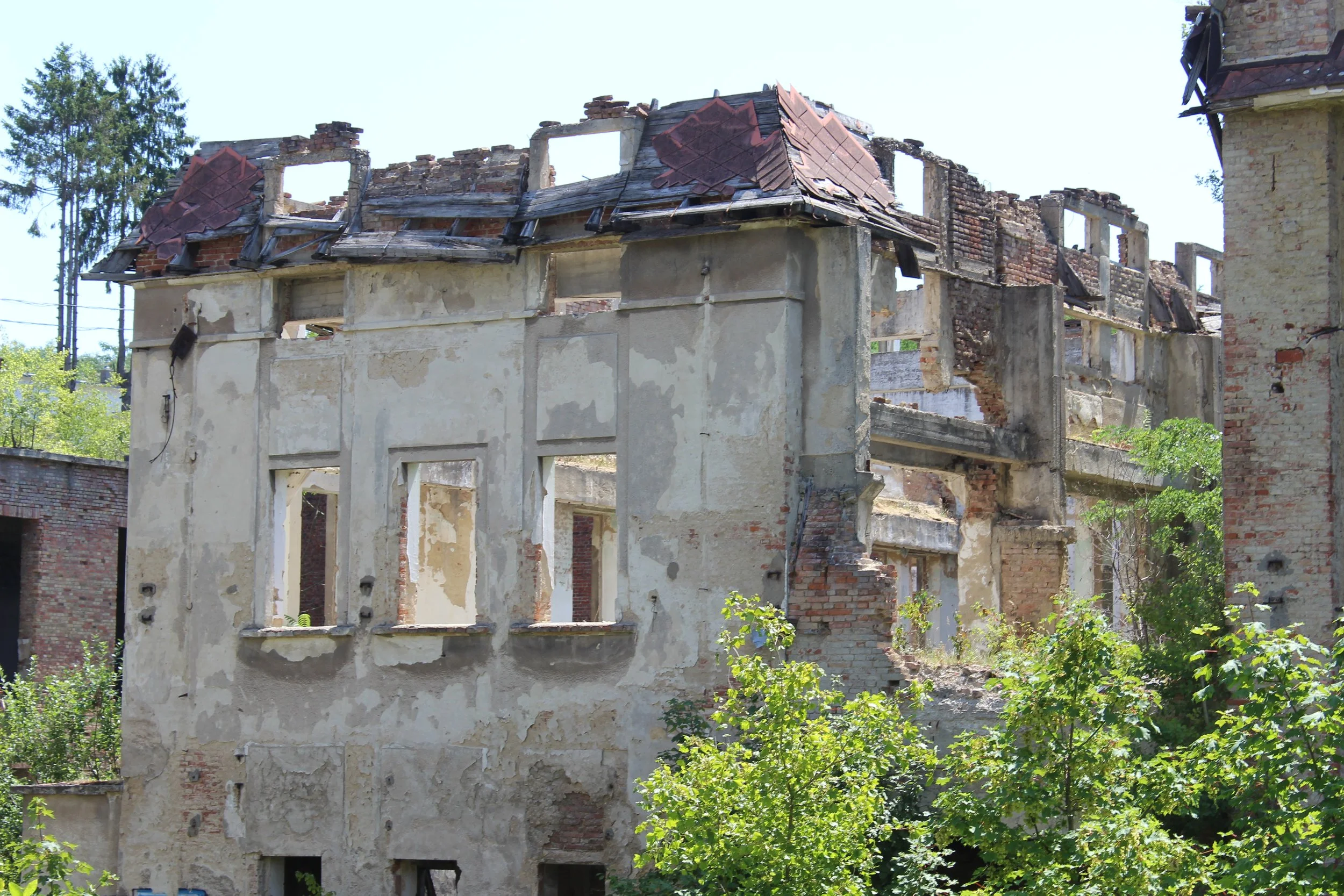 Verfallenes, zerstörtes Haus mit beschädigtem Dach und offenen Fenstern, umgeben von grünen Büschen und Bäumen.