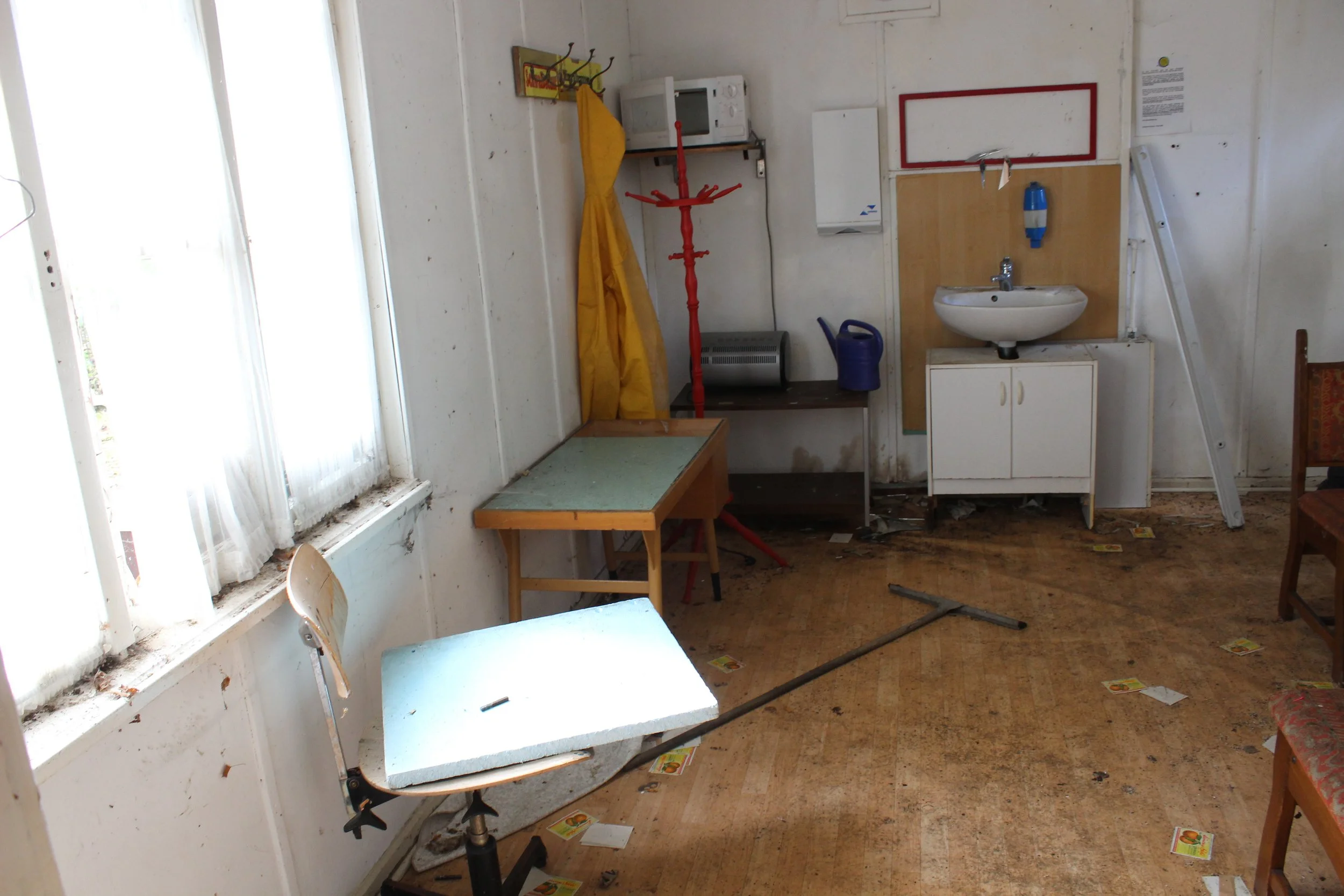 Ein verfallenes, schmutziges Zimmer mit einem zerstörten Schreibtisch, einer kaputten Holzstabel, einem Waschbecken an der Wand, einem Fenster mit schmutzigen Vorhängen und verstreuten Papierresten auf dem Boden.