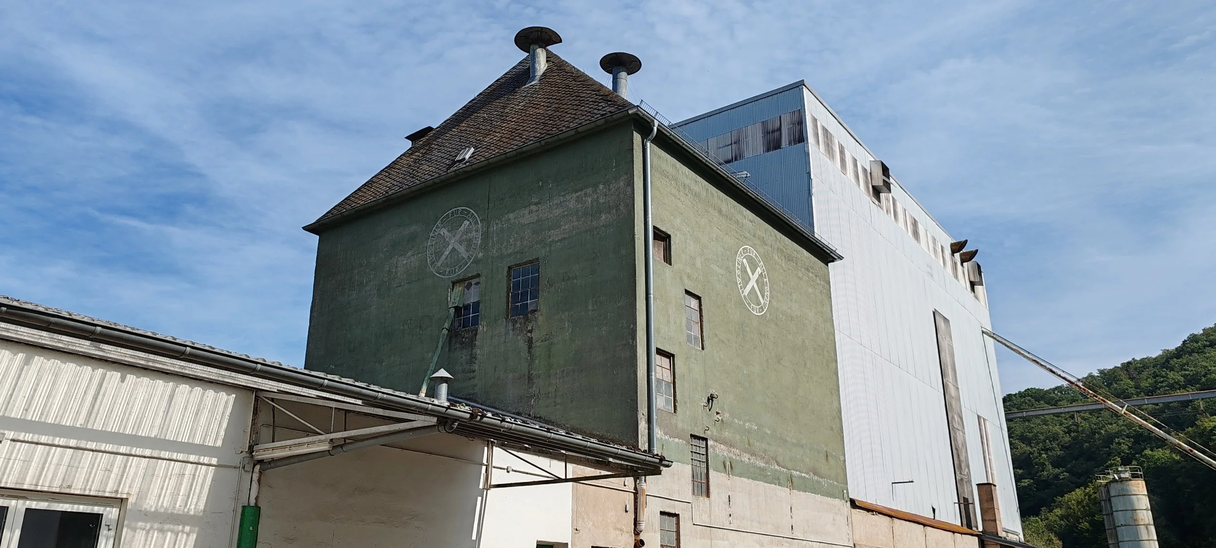 Altbau- und Industriegebäude mit grüner Außenfassade und moderner weißer Struktur, mit Status-Lampen und umliegendem Grünland, bei sonnigem Himmel
