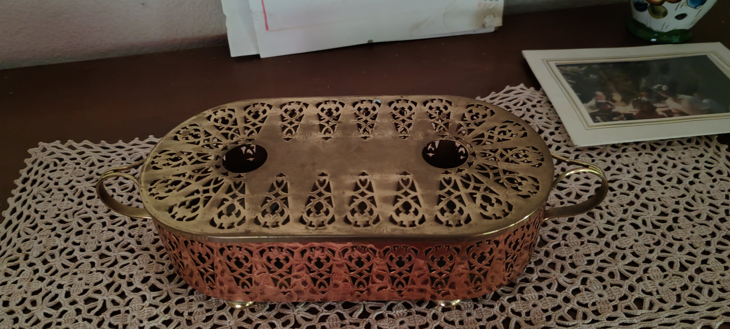 Messing-Keksdose mit filigranem Muster auf einem gehäkelten Tischdeckchen, daneben ein Familienfoto in Rahmen und eine grüne Dekoblume.