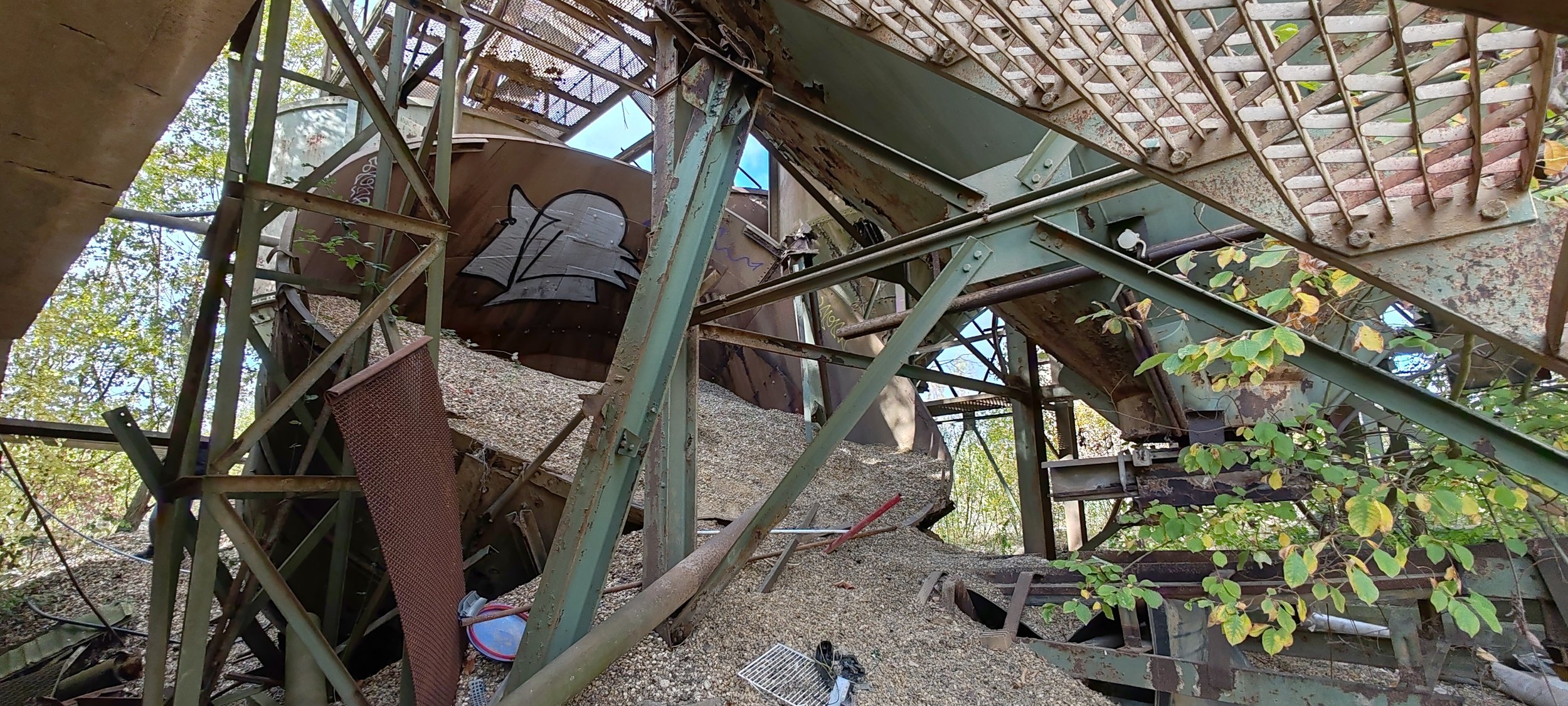 Verfallene, verrostete Industrieanlage mit Stahlstrukturen, Graffiti an einer Wand und überwachendes Laub von Bäumen