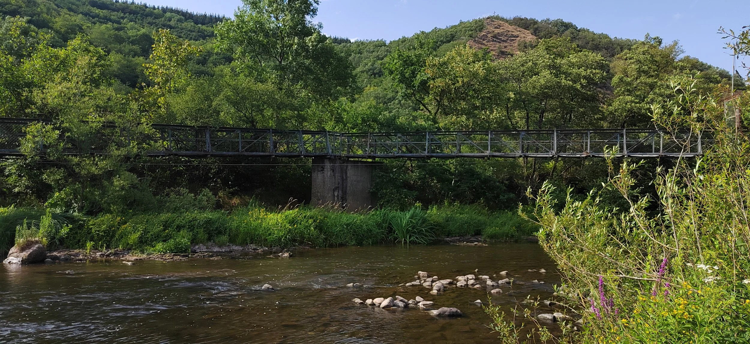 Blick auf einen Fluss mit kleinen Steinen, eine Brücke aus Metall und Beton, umgeben von üppigem Grün und Bäumen, mit einem bewaldeten Hügel im Hintergrund bei sonnigem Wetter.