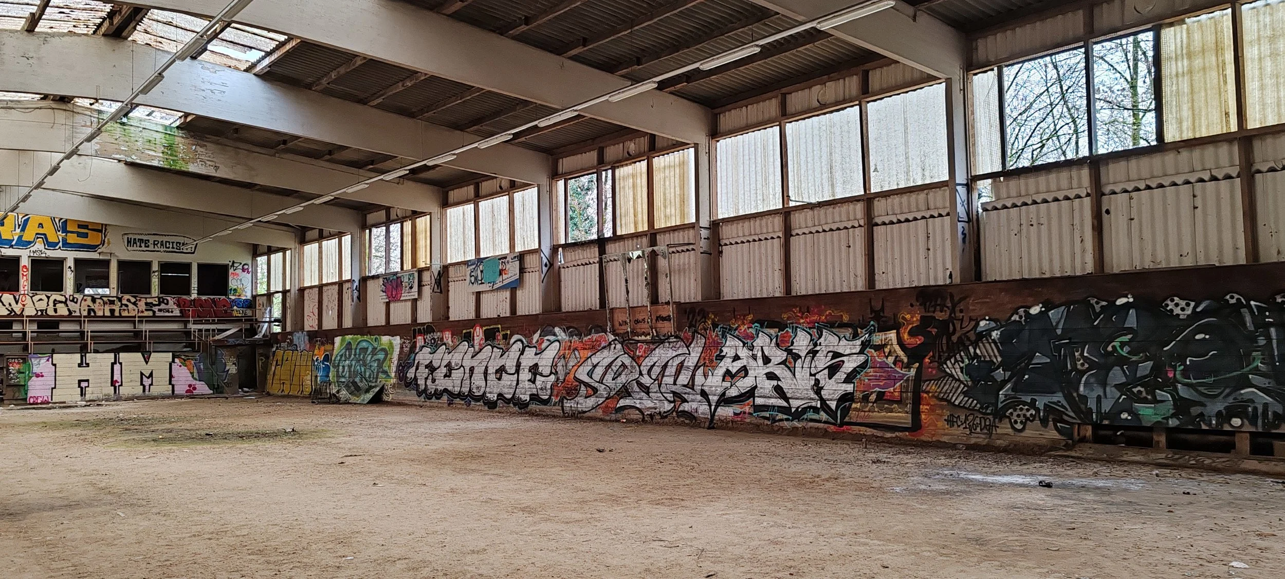 Innenraum eines verlassenen, verrosteten Sporthallen mit Graffiti auf den Wänden, Sandboden, hohe Fenster lassen natürliches Licht herein.