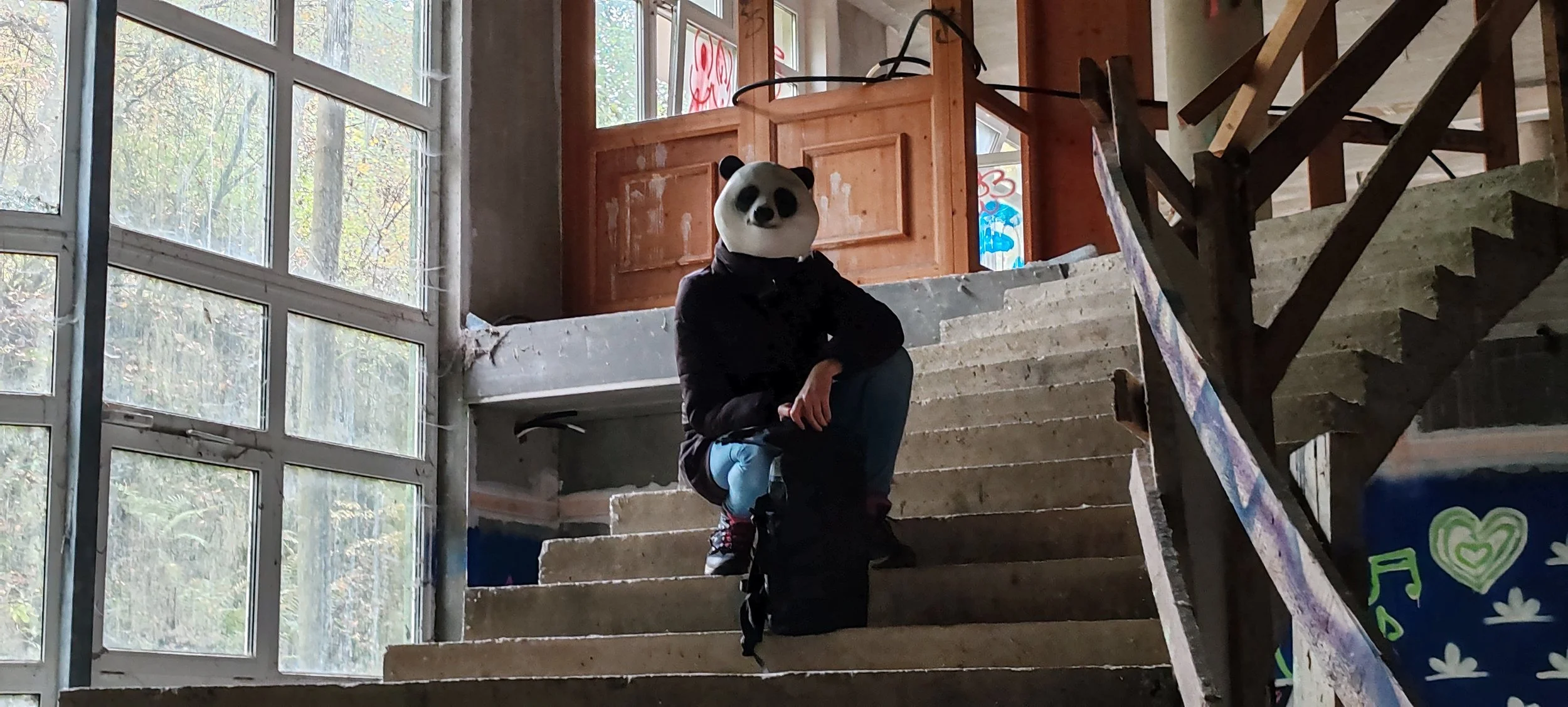 Person mit Panda-Maske, sitzend auf einer Treppe in einem ungewohnten Gebäude mit großen Fenstern