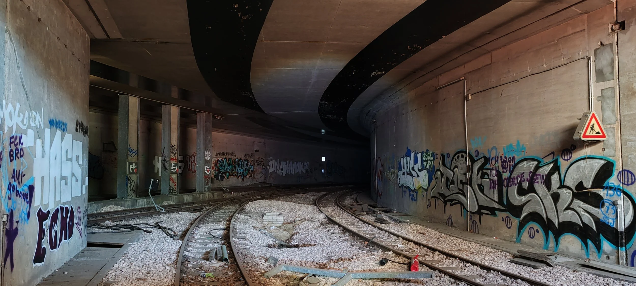 Ein Tunnel mit Graffiti an den Wänden und Schienen im Vordergrund, die im Bogen verlaufen. Es ist nicht sehr hell, und es gibt Baustellenschilder und -material auf dem Boden.