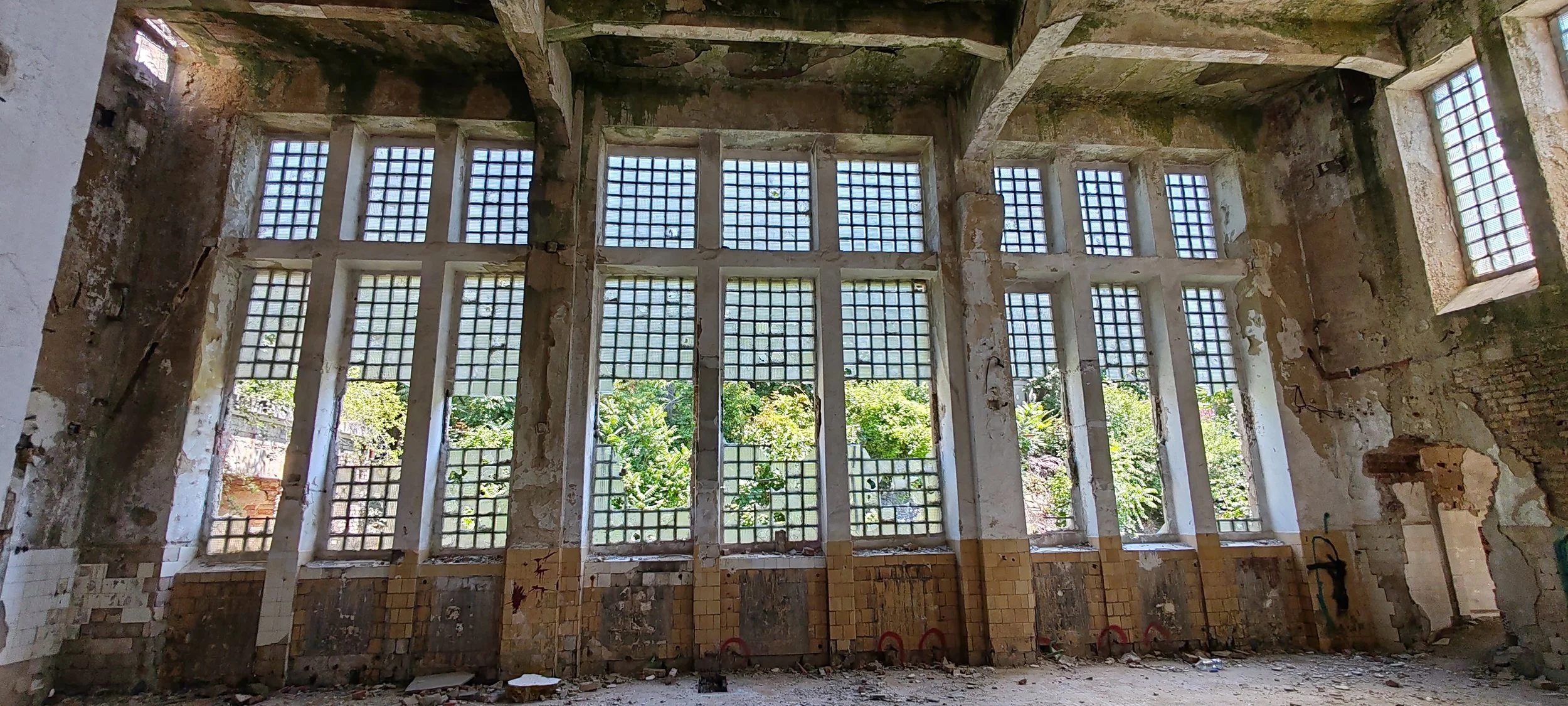 Ein verlassener, verfallener Raum mit großen Fensterflächen aus quadratischen Glasbausteinen, durch die Tageslicht einfällt, Blick nach draußen auf grüne Bflanzen und Bäume, sichtbare beschädigte Wände und Boden, alte Mauerreste, verfallene Struktur.