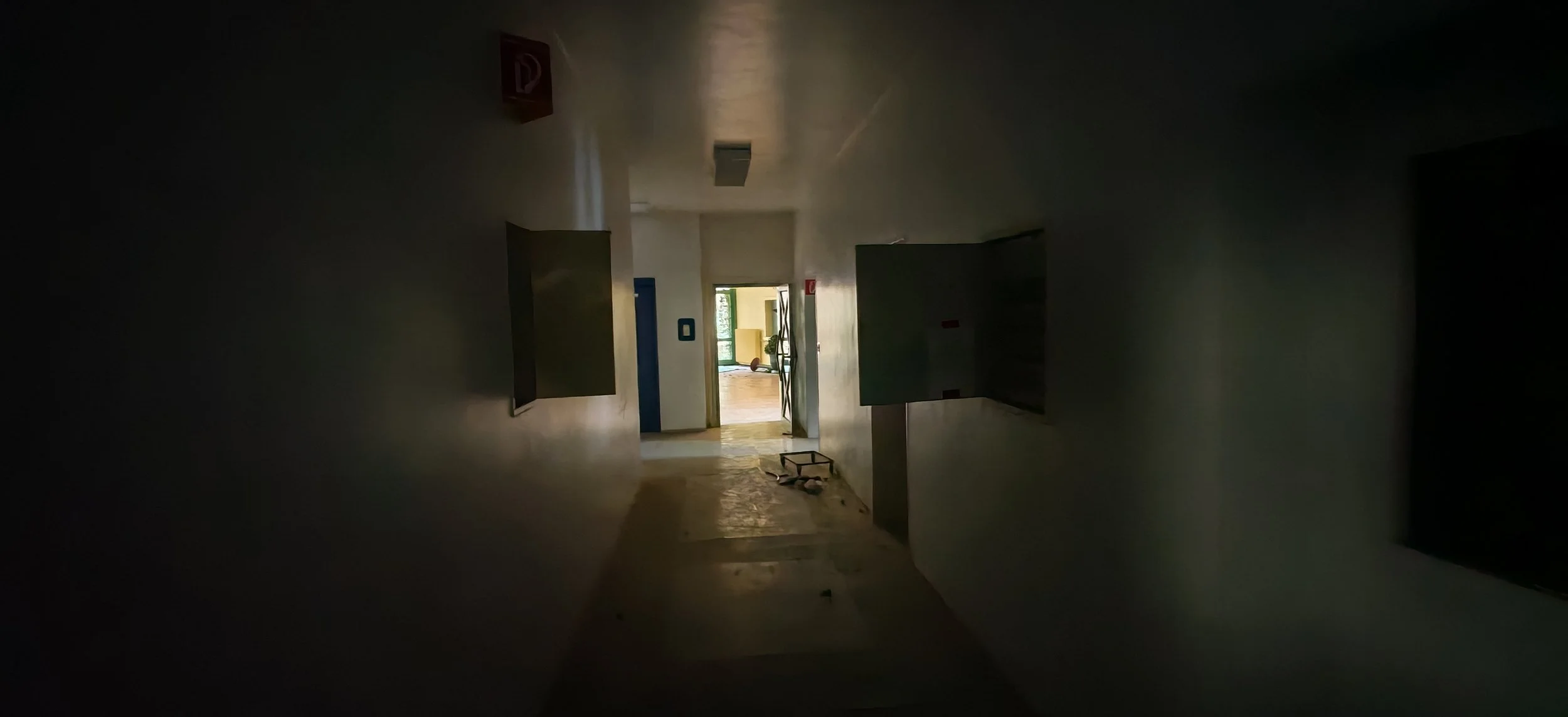 Ein dunkler Flur in einem verlassenen Gebäude mit einer offenen Tür, durch die man einen hell erleuchteten Raum mit Fenstern und einem Kugelobjekt sehen kann.